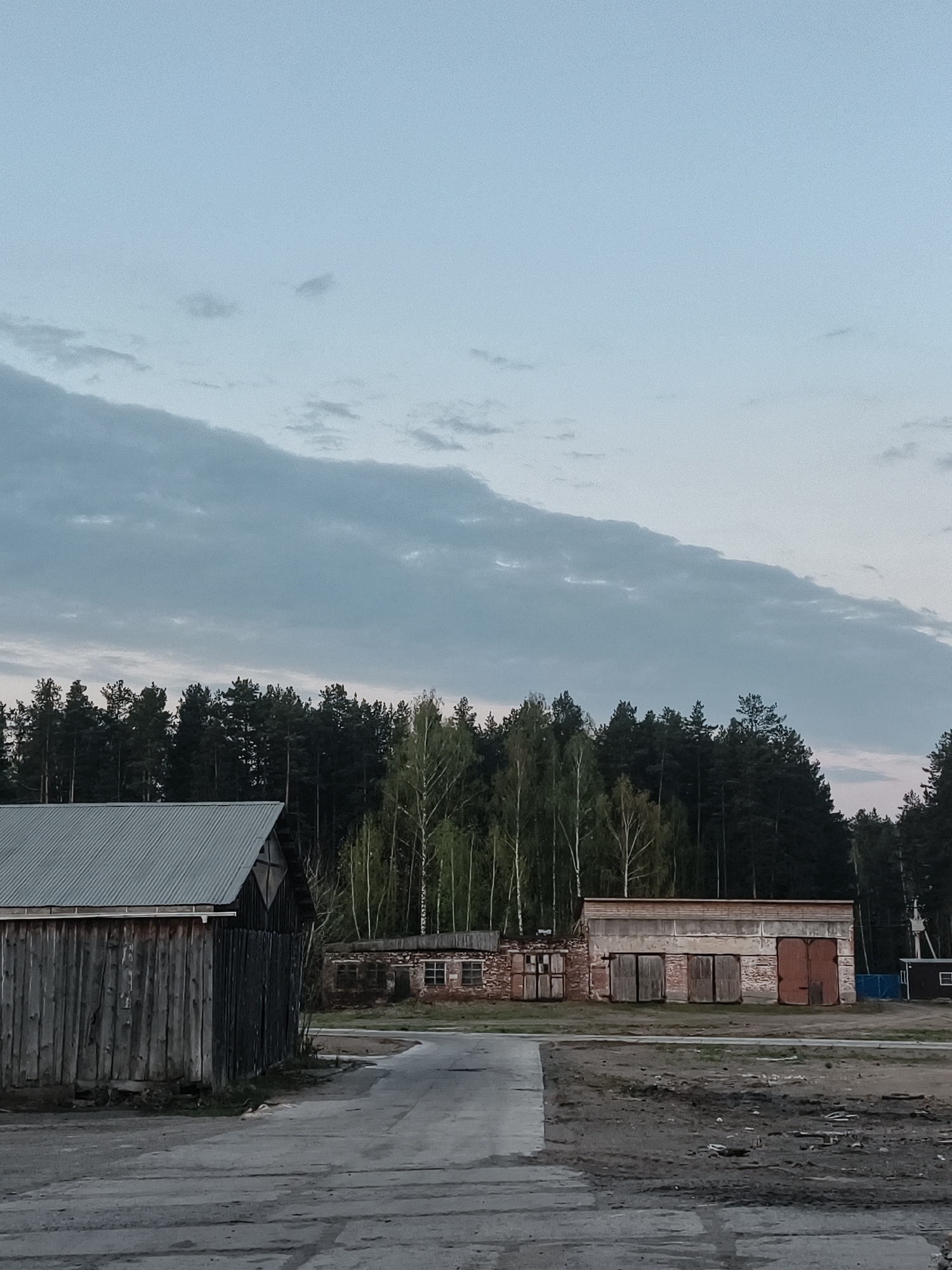 The image depicts a rural or industrial scene in Noviy, Russia, under an overcast sky. In the foreground, a weathered wooden barn-like structure stands to the left of a paved road or area. Directly ahead is a brick building with multiple closed doors, possibly a garage or storage facility. A line of trees, including birch and evergreens, forms a backdrop. The setting appears quiet and possibly abandoned.