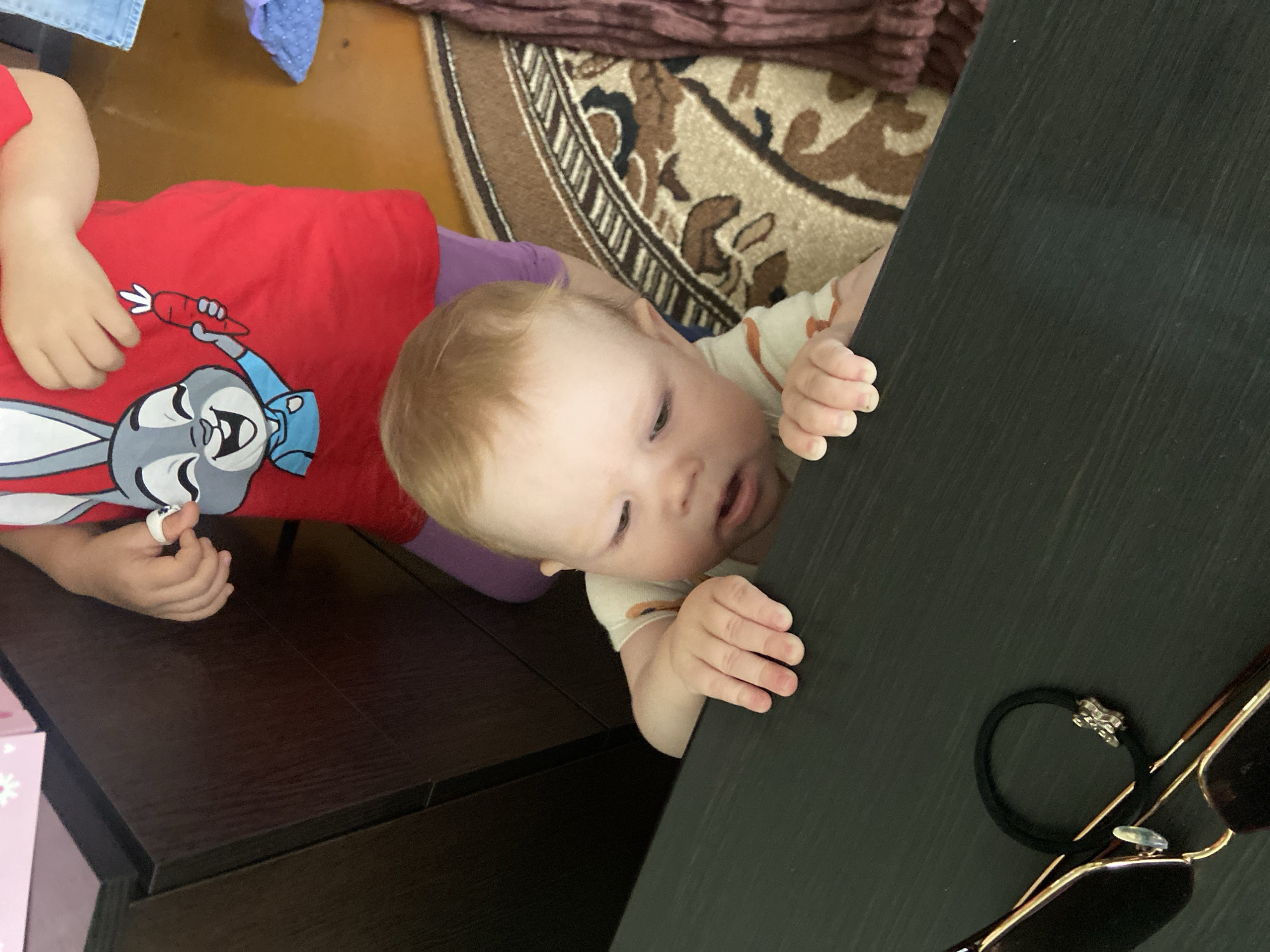 A high-angle shot shows a baby with blonde hair, wearing a light-colored shirt with stripes, grasping the edge of a dark-colored table. To the left of the baby, a child wearing a red shirt with a Bugs Bunny graphic is also near the table. Sunglasses and a black hair tie sit on the table's surface. A patterned rug is partially visible in the background. The location is not discernible from the image.