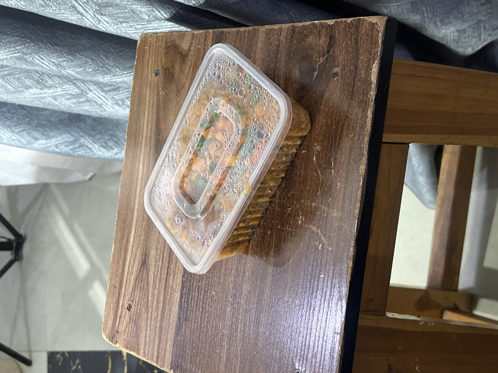 The image shows a clear plastic rectangular food container filled with a meal, placed on a rustic wooden surface. The food inside appears to be rice or pasta with vegetables and possibly some protein, with a reddish-brown sauce. Condensation is visible on the inside of the container lid. The wooden surface has a worn, weathered appearance with visible grain patterns and some surface imperfections. In the background, a grey, textured fabric is draped, and parts of a wooden chair or table structure are visible. The lighting suggests an indoor setting, possibly in the daytime, given the diffused light. The overall scene conveys a simple, casual presentation of a meal. There is no visible text in the image. The location context of Gura Top, Nigeria, is provided but not visually evident from the image itself.