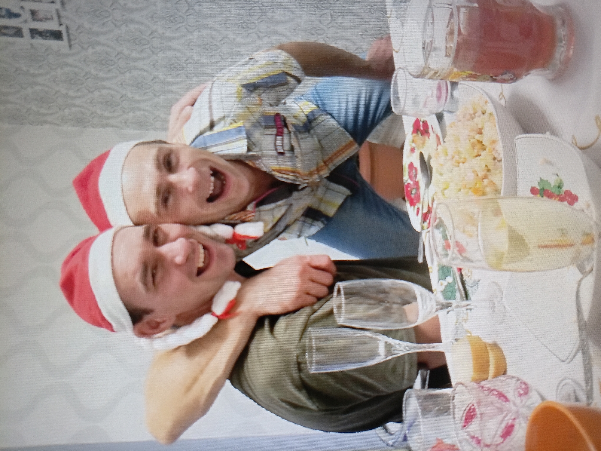 Two men are pictured indoors, likely celebrating a holiday as they are both wearing red and white Santa hats. They are positioned closely, with one man's arm around the other's shoulders. Both men have open mouths and appear to be laughing or speaking animatedly. They are seated at a table covered with a white tablecloth, upon which several glasses and a bowl of food are visible. The man on the left wears a dark green t-shirt, while the man on the right is dressed in a plaid shirt. The background consists of a patterned wallpaper and what appears to be a shelf with framed pictures. The image is slightly blurry, suggesting it was taken with a mobile phone camera or under low-light conditions. The overall impression is a candid and joyful moment captured during a social gathering.