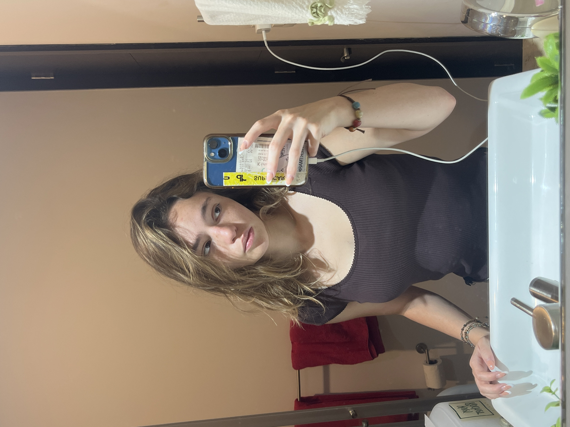 A person is taking a selfie in a bathroom mirror. The person has light brown hair and is wearing a dark top with a light-colored trim around the neckline. The person is holding a blue smartphone, which is plugged into a white charging cable. The phone has a ticket or receipt affixed to the back. The background shows a beige wall, a black cabinet, a white sink, a red towel on a rack, and bathroom fixtures. No identifying information is available to determine the location is San Bernardino Tlaxcalancingo, Mexico.