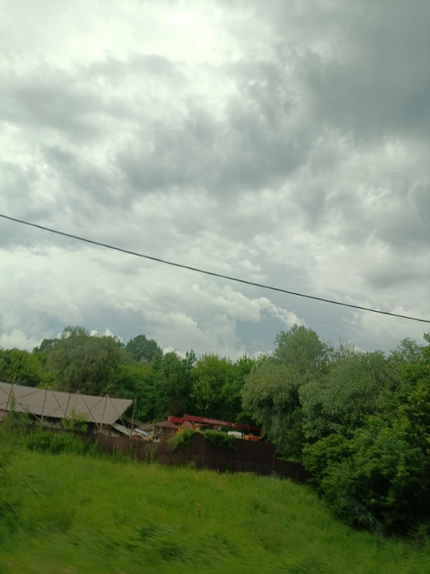 The image depicts an outdoor scene with a cloudy sky dominating the upper portion. Below the sky, a dark, thin wire stretches horizontally across the frame. In the middle ground, a dense line of green trees forms a lush backdrop. A low-lying structure with a light-colored roof and exposed wooden beams is partially visible on the left, situated behind a dark fence. To the right of this structure, there appears to be a red object, possibly part of a vehicle or equipment, and a small gazebo. The foreground consists of a bright green, grassy slope that appears to be moving or blurred, suggesting the photo was taken from a moving vehicle. The overall impression is a rural or semi-rural setting, possibly on the outskirts of Saratovskaya, Russia, under overcast conditions.