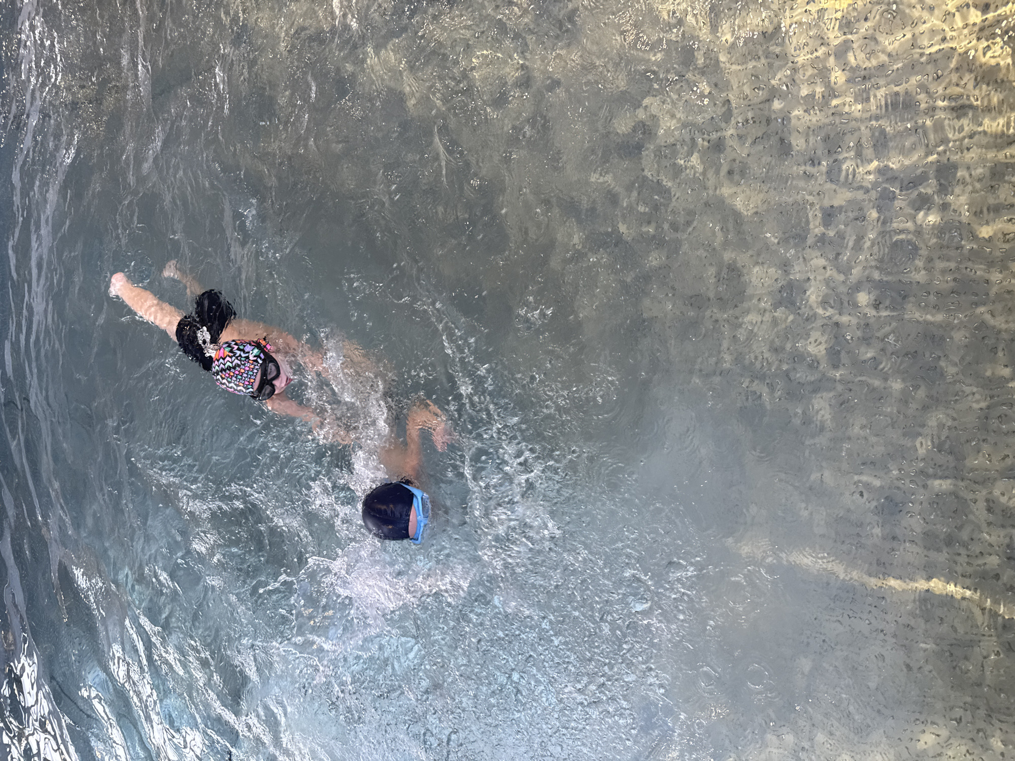 In the city of Sovkhoz imeni Lenina, Russia, two individuals are swimming in a pool. One person, wearing a colorful swim cap and goggles, is in the upper right portion of the image, swimming on their front with their legs kicking. To the left, another person is visible, also swimming on their front, wearing a dark swim cap and blue goggles. They are closer to the foreground, with water splashing around them. The pool water is clear and appears to be illuminated from above, with light reflecting off the surface. The bottom of the pool is visible in some areas, appearing to be tiled or textured.
