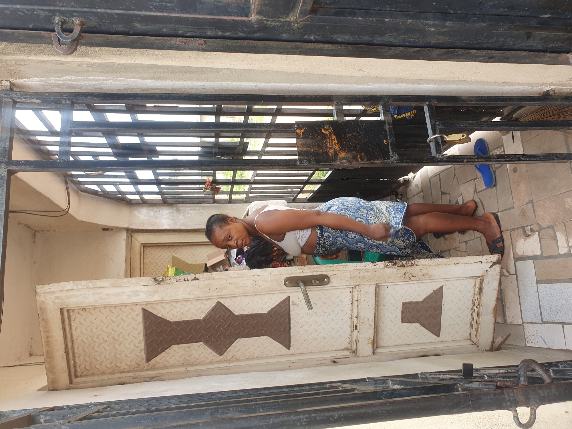 A medium shot captures a woman standing in a doorway in Lokoja, Nigeria. She is wearing a white tank top and a blue and white patterned skirt, with dark sandals on her feet. She is looking towards the camera with a neutral expression. The doorway is framed by a black metal gate, and the door itself is white with brown diamond-shaped accents. The floor is tiled with a geometric pattern. In the background, another door is visible, and stacks of boxes and fabric are piled near the entrance. Sunlight streams through the metal gate, creating strong shadows.