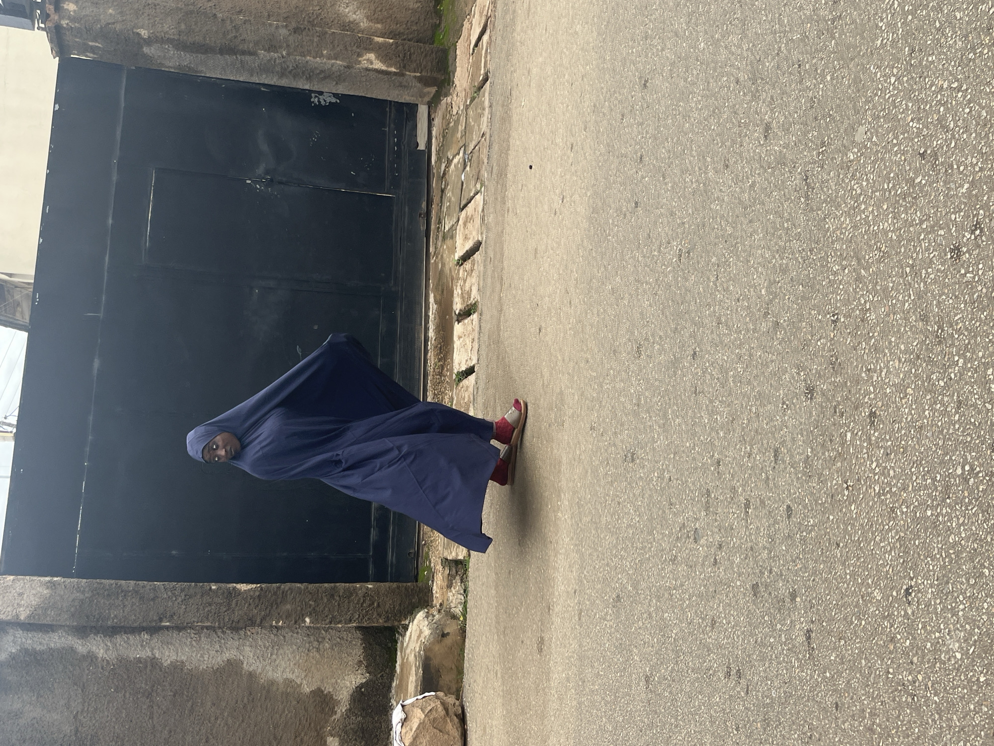 A person, who appears to be female, is standing on an asphalt road in front of a large black gate. She is wearing a navy blue abaya that covers her body and head, with only her face and feet visible. She is also wearing red sandals with white socks. The gate is made of metal and has a textured surface. To the left of the gate is a rough, grey concrete wall. The ground in front of the gate is paved with stone slabs, some of which have small plants growing between them. The road surface is asphalt, with small pebbles visible. The lighting suggests it is daytime, possibly late morning or early afternoon, with the sun casting shadows. The scene appears to be in an urban or semi-urban area. No text is visible in the image. The person's facial expression is not clearly discernible.