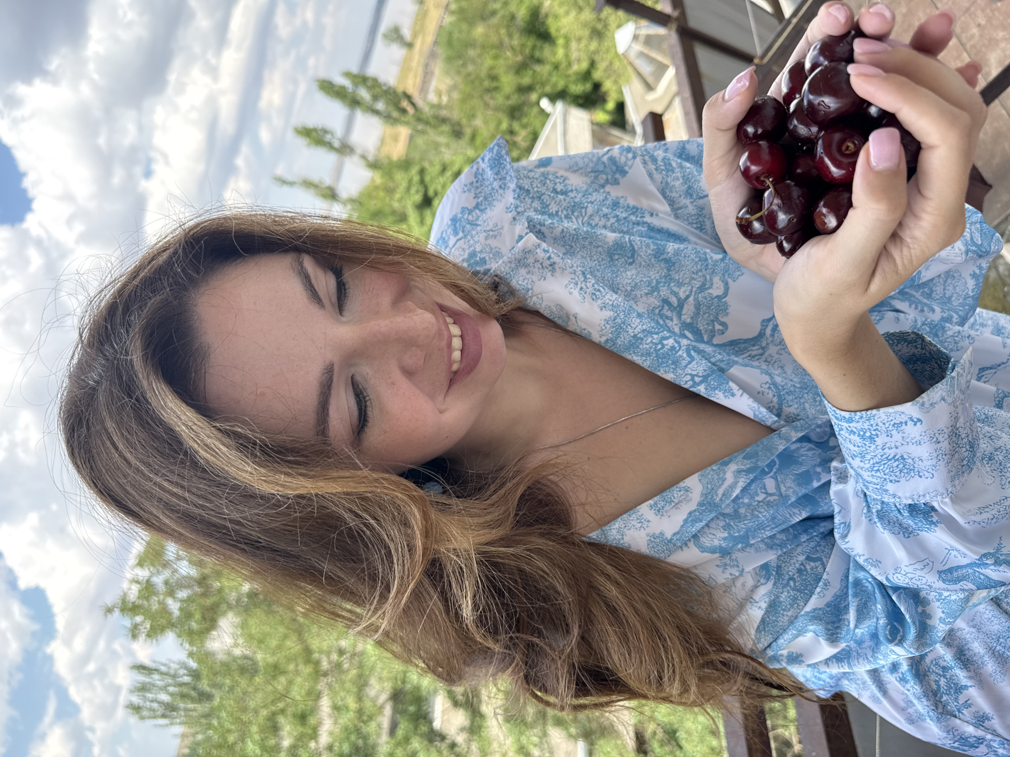 A young woman is holding a pile of dark red cherries in her cupped hands. She is wearing a blue and white patterned dress. Her eyes are closed, and she has a wide smile on her face, suggesting a moment of joy or pleasure. Her long, light brown hair frames her face. In the background, there are green trees and a cloudy sky, indicating an outdoor setting during the daytime. The presence of trees and a railing suggests a balcony or an elevated outdoor space. The overall scene conveys a sense of happiness and enjoyment of nature and fresh fruit.