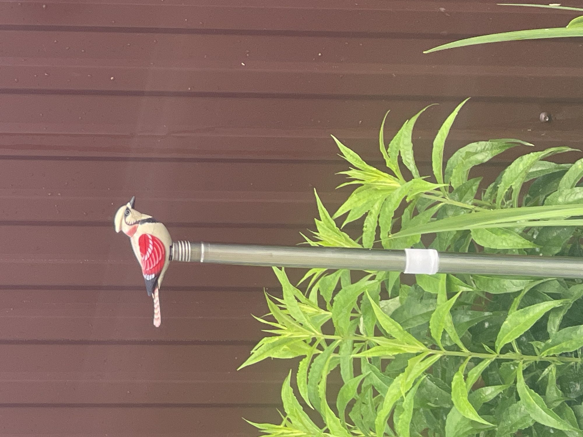A decorative bird figurine is perched atop a metal pole in front of a brown, corrugated metal fence. The bird is predominantly white with red wing accents and black and blue markings. Green foliage surrounds the base of the pole. The image was taken in Mesyagutovo, Russia.