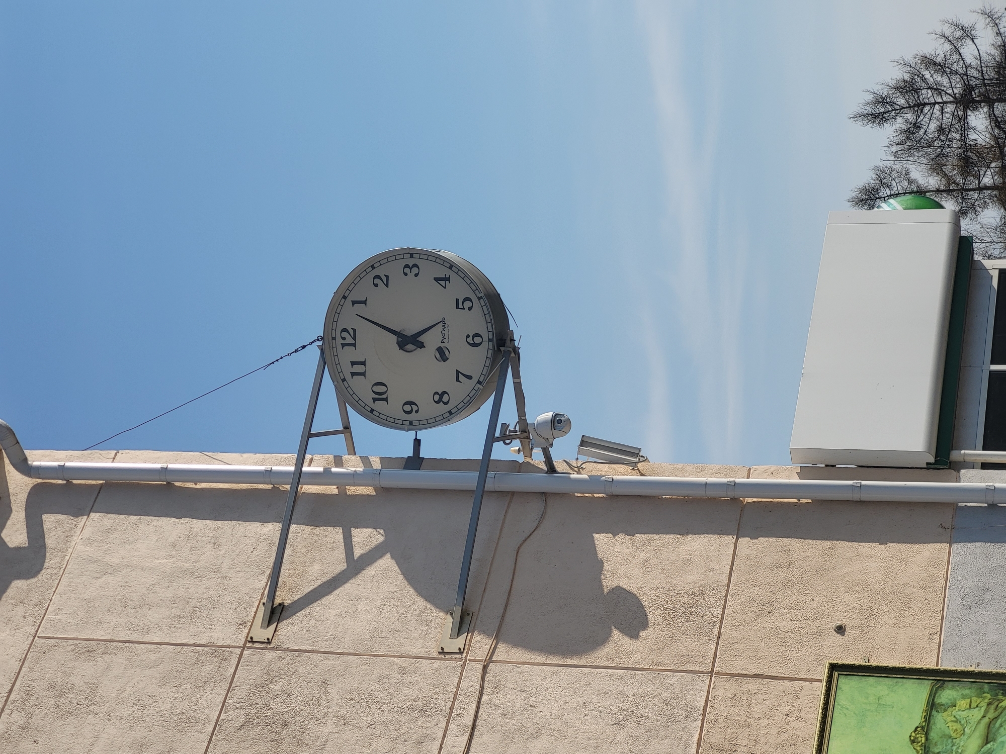 The image shows a clock on a building in Chaikovsky, Russia. The clock has a white face with black numbers and hands, and it is mounted on a metal stand. A security camera is mounted near the clock. The building has a textured beige exterior and white trim. A white box and tree branches are visible on the roof. The sky is blue with some clouds.