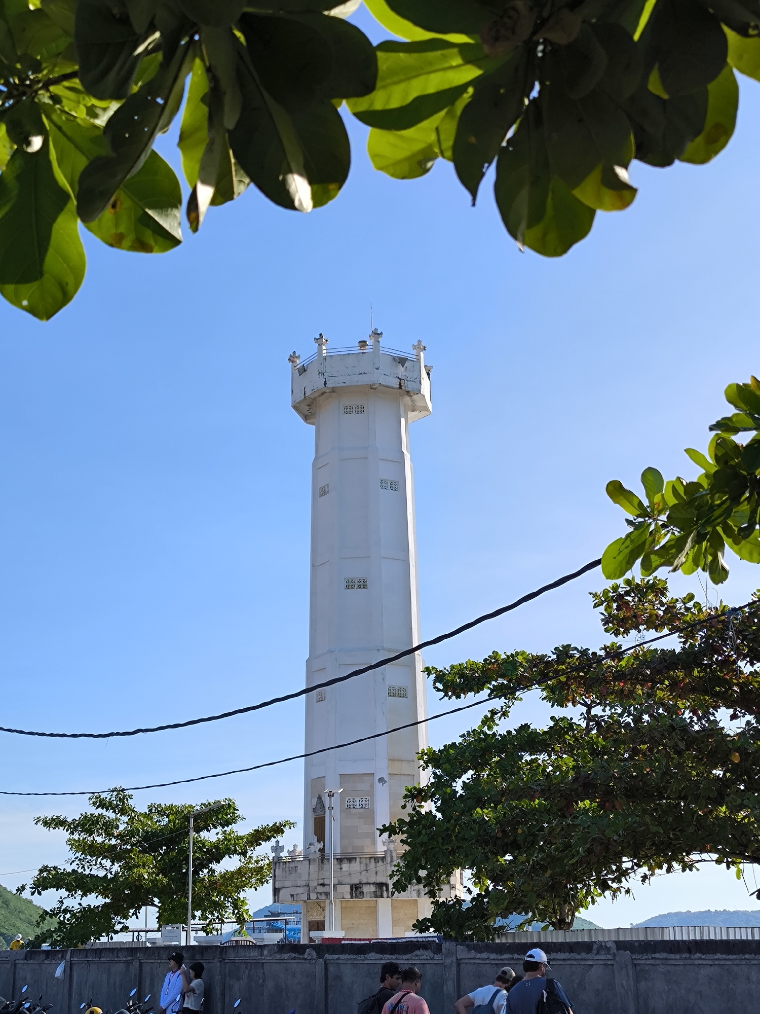 The image depicts a tall, white lighthouse with a hexagonal shape, located in Nusa Penida, Indonesia. The structure has multiple levels and is topped with a decorative railing. The sky is clear and blue. The scene is framed by tree branches and leaves at the top and sides. In the foreground, a concrete wall with people and parked motorbikes is visible. The view extends towards the horizon with hints of hills in the distance. Power lines run across the view. The image appears to have been taken during the day in bright sunlight.