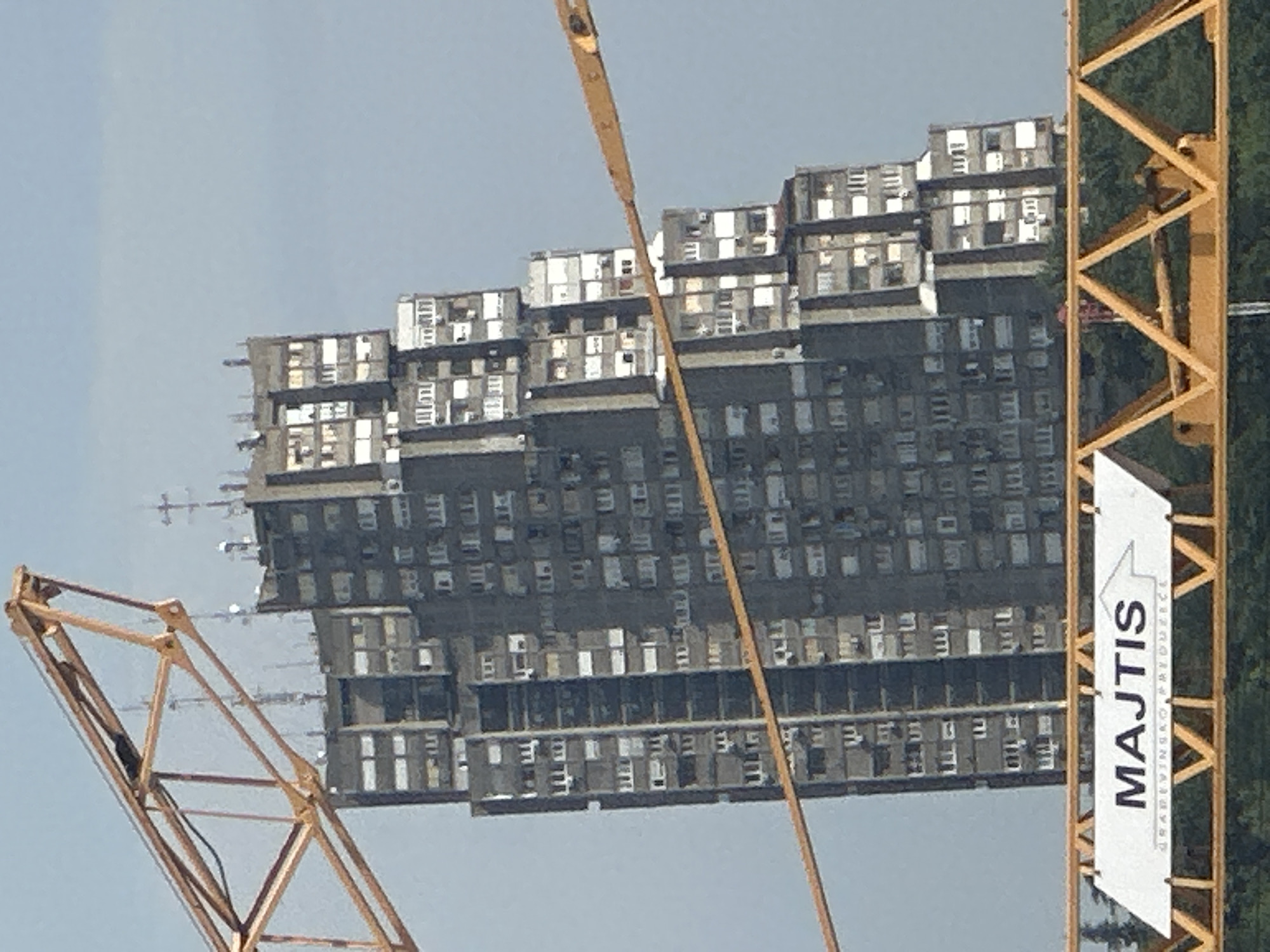 The image depicts a tall, Brutalist-style apartment building under construction or renovation, seen from a low angle, possibly from a crane or a similar elevated position. The building is characterized by its repetitive, modular design, with numerous windows and balconies. The perspective is distorted, making the building appear to stretch horizontally across the frame.
A yellow construction crane is prominently featured, dominating the right side of the image. Part of the crane's structure, including its lattice boom, is visible. A white banner with text is attached to the crane's vertical support. The text on the banner reads "MAJTIS" in large, bold, black letters, with smaller text below it.
The sky is a hazy, light blue, suggesting an overcast or dusty day. The overall scene conveys a sense of urban development or reconstruction. The time of day is likely daytime, given the illumination. The location cues of Belgrade, Serbia, are provided, which aligns with the architectural style and the presence of construction equipment. No people are visible in the frame, focusing solely on the building and the crane. The image has a somewhat surreal quality due to the distorted perspective and the starkness of the architecture against the sky.