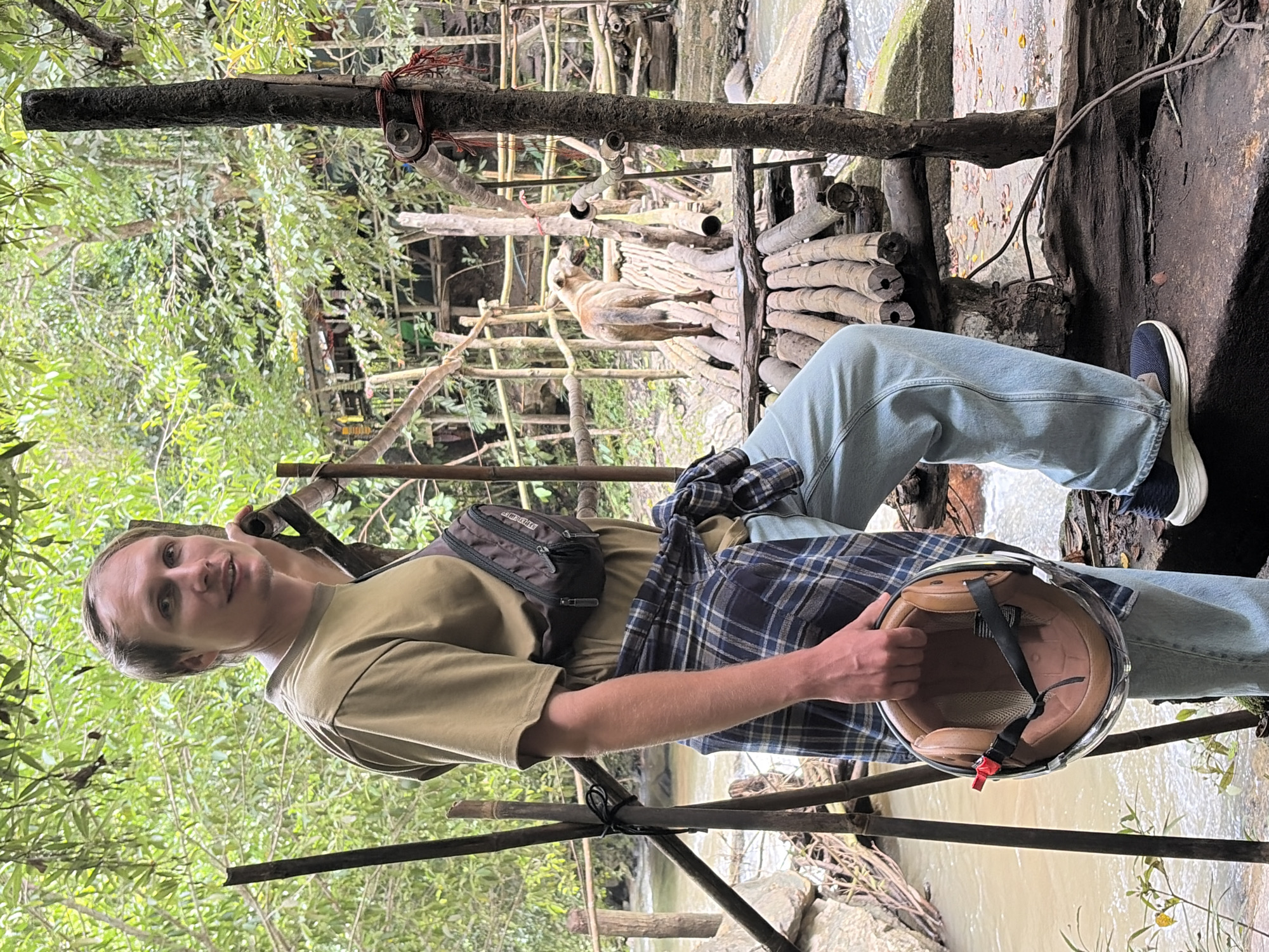 A man stands in front of a natural scene in Ban Huai Takhrai, Thailand. He is wearing a tan t-shirt, a blue and black plaid shirt tied around his waist, light blue jeans, and dark sneakers. He holds a brown and black motorcycle helmet in his right hand. A small brown dog stands on a wooden bridge in the background, facing away from the camera. The environment consists of lush green trees and foliage, with a body of water flowing through rocks. A rustic wooden structure, possibly a bridge or walkway, is visible in the mid-ground. The lighting suggests it is daytime, with diffused natural light.