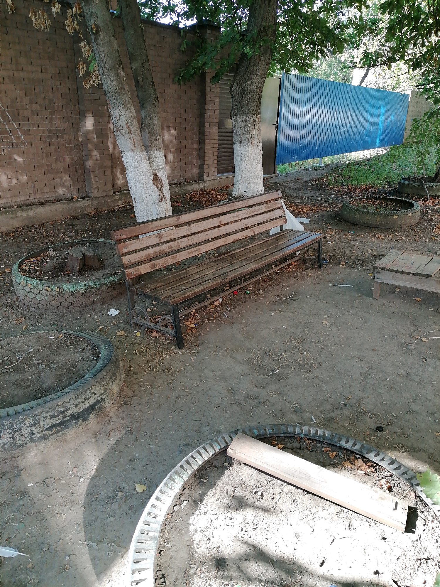 The image depicts an outdoor area in Rostov-on-Don, Russia, possibly a courtyard or a section of a park, characterized by a weathered wooden bench as the central subject. The bench, made of slats, is positioned on a dirt ground covered with scattered dry leaves. To the left of the bench, two trees with whitewashed lower trunks stand against a brick wall. In the background, a tall, blue corrugated metal fence is visible, separating the area from what appears to be a street or another outdoor space. Several large, old tires are scattered around the ground, some serving as planters or decorative elements, with one tire in the foreground containing a wooden plank. The overall scene suggests an unkempt but functional outdoor space.