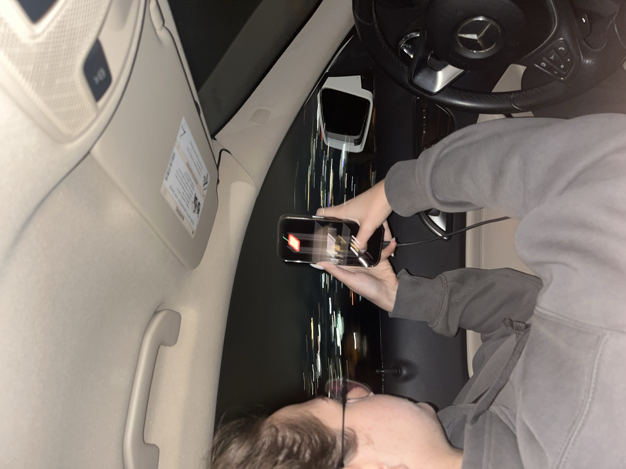 A person is in the driver's seat of a Mercedes-Benz vehicle. They are holding a smartphone and appearing to take a photograph of the street outside. The streetlights outside are blurred, suggesting movement. The location cannot be determined to be Blagoevgrad, Bulgaria from the visual information provided. The person is wearing a grey hoodie.