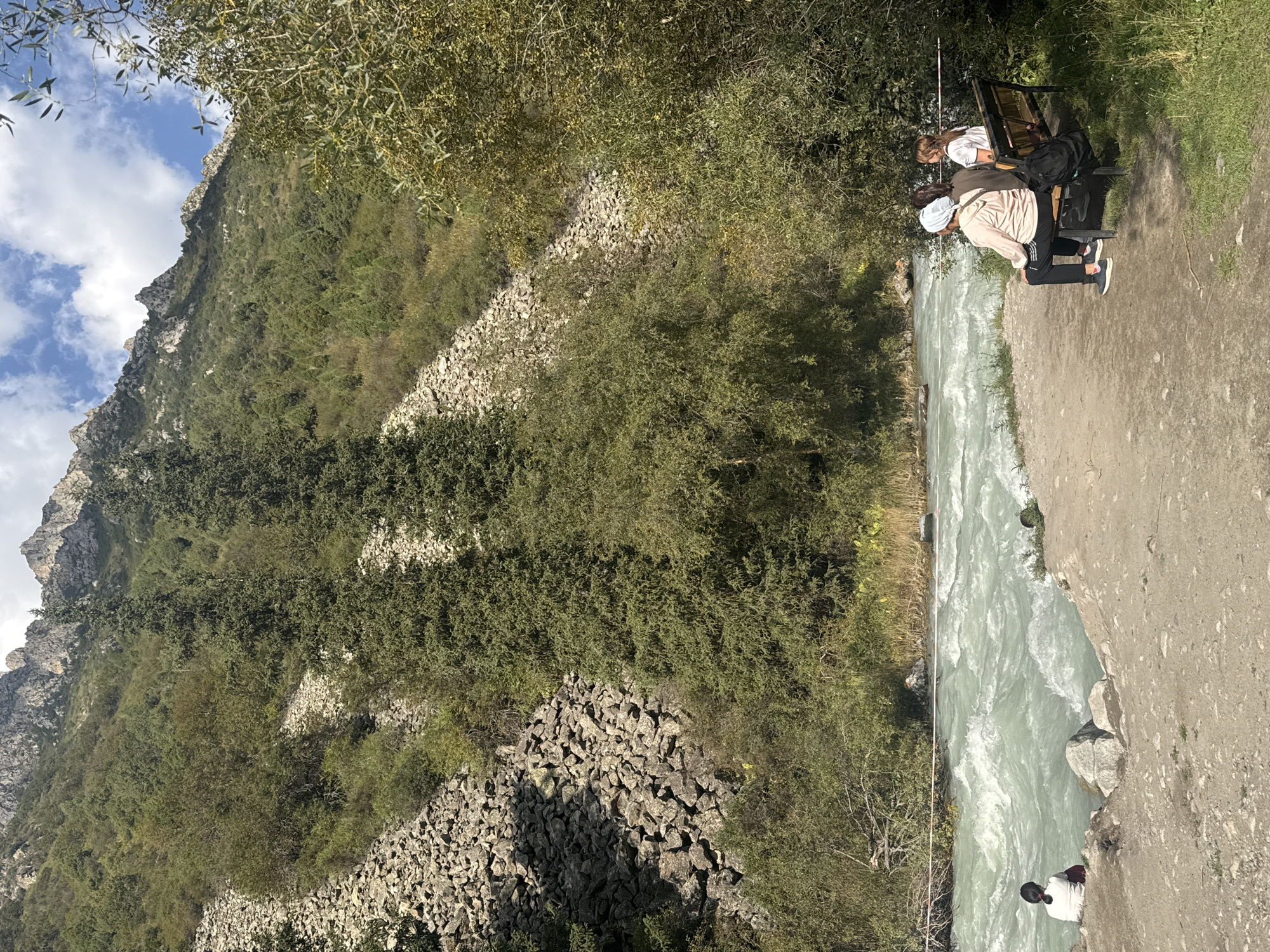 A wide shot captures a turbulent, turquoise river flowing through a mountainous landscape in an unidentified location in Kyrgyzstan. Steep, rocky slopes covered in dense green vegetation, including coniferous trees and shrubs, rise dramatically on either side of the river. The sky above is partly cloudy, with patches of blue showing through. In the foreground, a gravel path leads to the riverbank. On the path, three individuals are seated on a wooden bench, facing the river. Two of them are women with their backs to the camera; one wears a light pink hoodie and black athletic pants, the other a dark shirt. A third woman, partially obscured, is also on the bench. To the left of the bench, near the river's edge, a single person is sitting with their back to the camera, wearing a white shirt and dark bottoms. The river appears fast-moving with white water rapids.