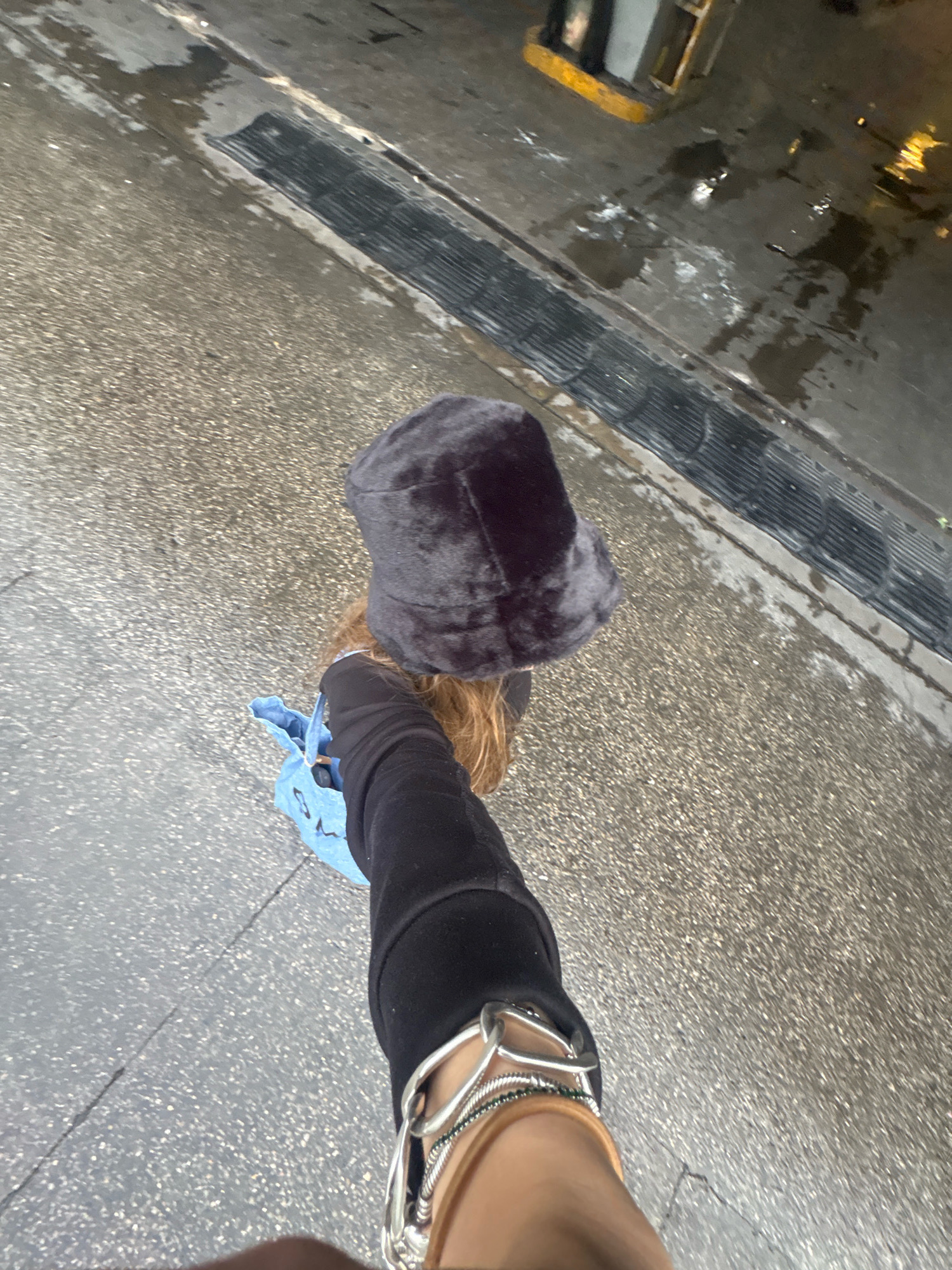 A person is viewed from a high-angle, first-person perspective, walking on a wet, textured pavement. They are wearing a dark, fuzzy bucket hat, a black long-sleeved garment, and blue denim shorts. The person is also carrying a light blue bag with a cartoon-like design. Several silver bracelets are visible on their right wrist, along with what appears to be a tan watchband. In the background, a dark metal grate runs parallel to the pavement, and the ground is marked with various dark wet patches, suggesting recent rain. Further back, a bright yellow curb and dimly lit structures are visible, hinting at a semi-urban or commercial environment. The overall scene suggests an overcast day in Mexico City, Mexico.