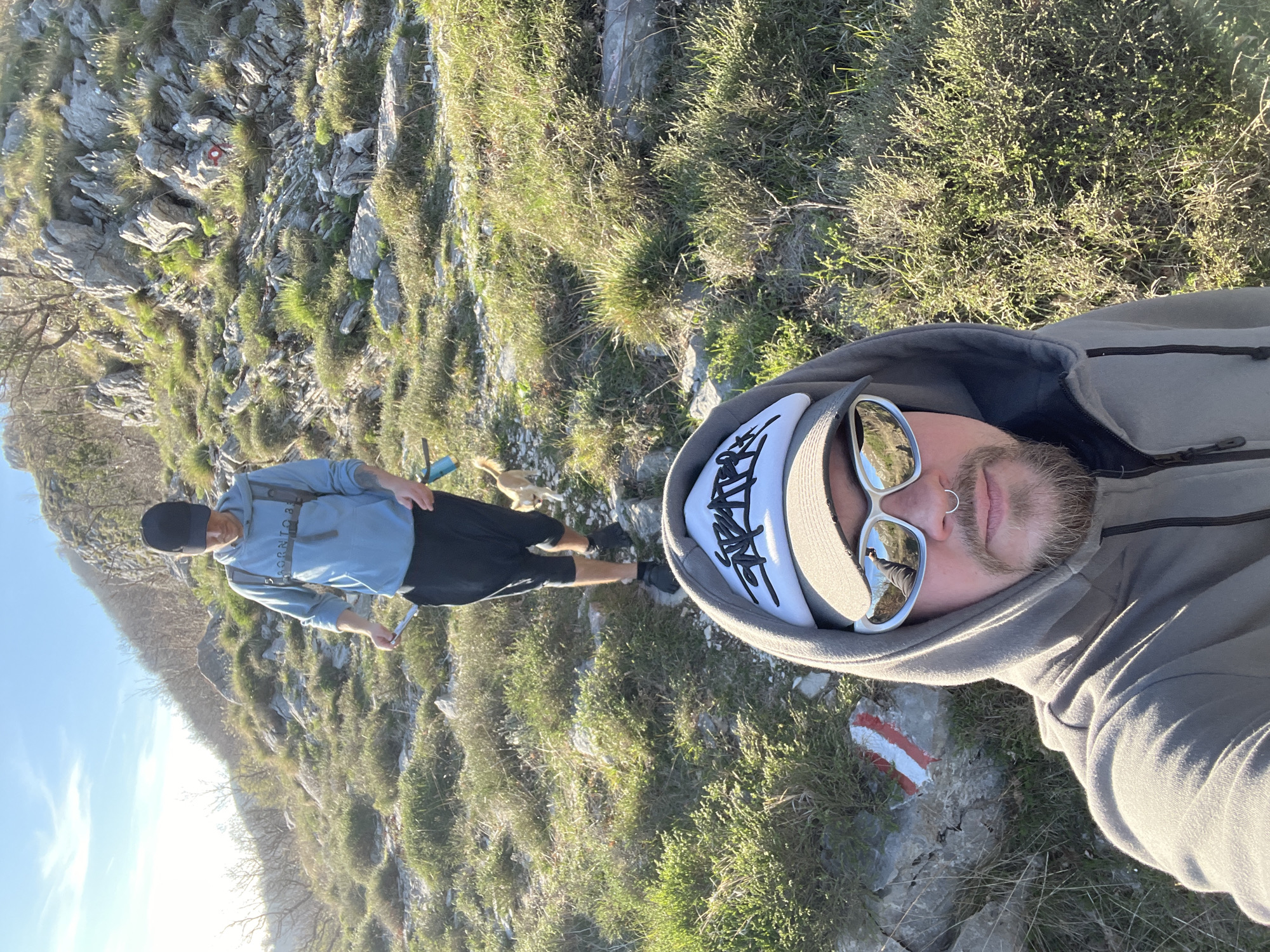 The image shows a selfie taken on a hillside, presumably in Dolovi, Montenegro. Two men are visible. The foreground features a man in a grey hoodie with a white cap and sunglasses. His face is prominent, capturing details such as a nose ring and beard. In the background, a second man is seen wearing a blue sweater, black shorts, and a black cap. He is walking on a rocky, grassy path, and a small dog is walking near him. The environment consists of rocks, grass, and low shrubs typical of a mountainous terrain. A red and white trail marker is also visible on a rock near the person taking the selfie. The sky is visible in the top left corner.