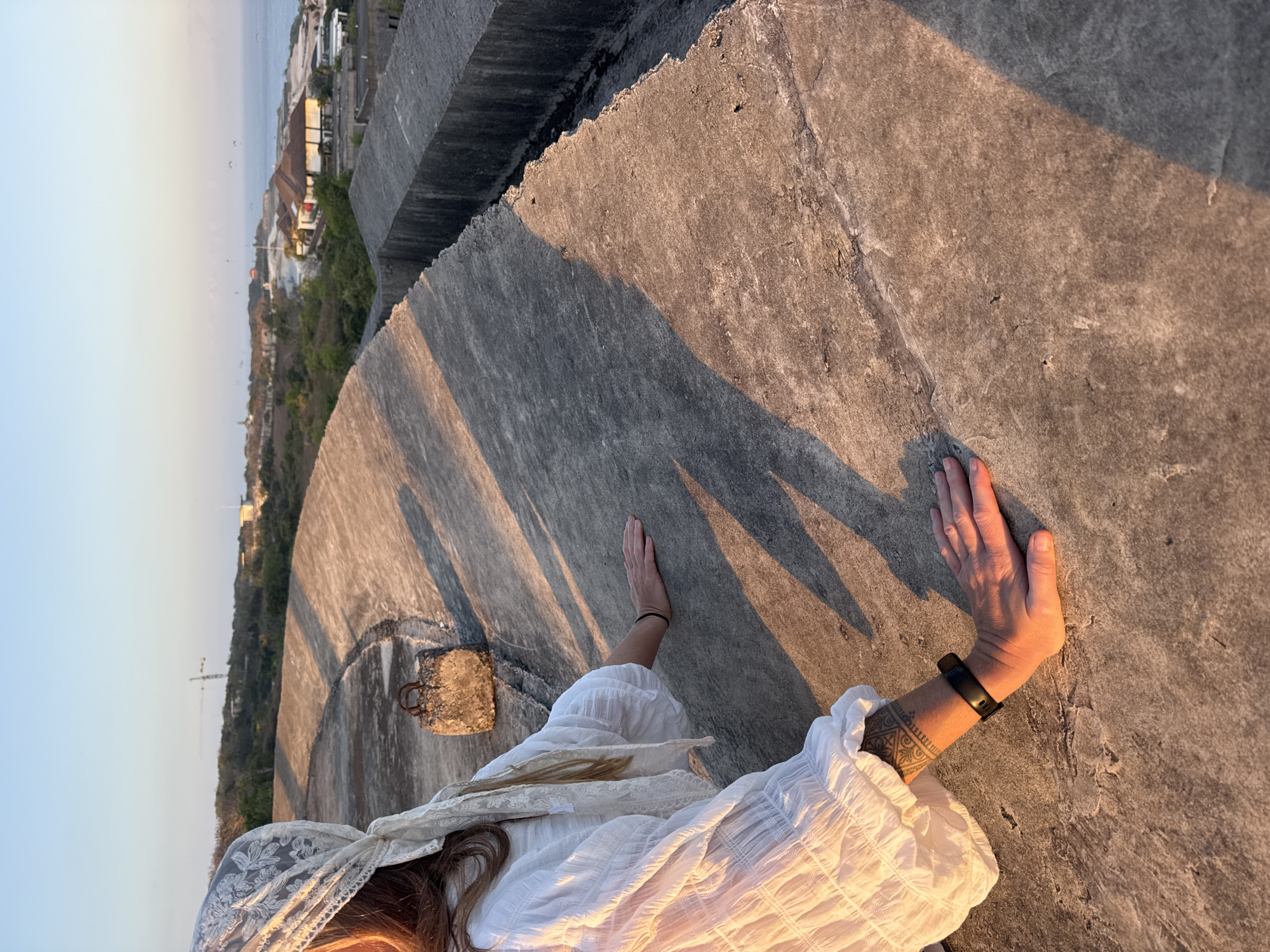 A person wearing a white, gauze-like top and a white lace headscarf is sitting on a concrete structure. The person's left hand is resting on the concrete, and their right arm is bent with their hand on their lap. A black fitness tracker is on their left wrist, and a tattoo is visible on their forearm. The background shows a coastal view with buildings, trees, and the ocean. The sun is low in the sky, casting long shadows on the concrete. The location appears to be a viewpoint overlooking a coastal area in Kuta Selatan, Indonesia, likely a historical site or overlook.