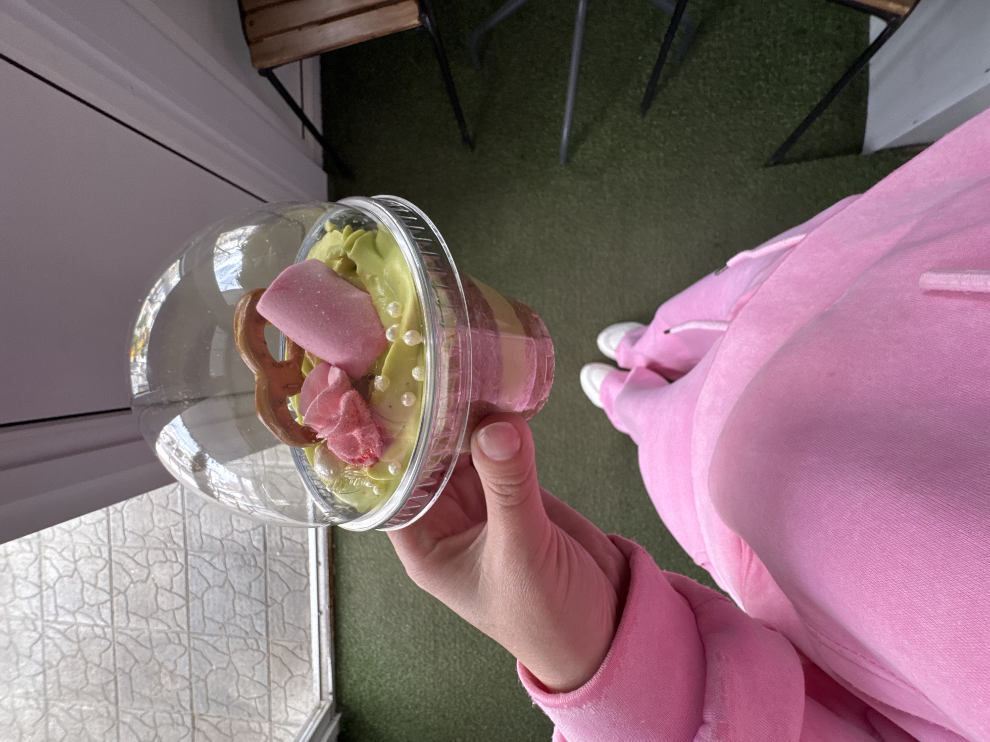 A person is captured in a high-angle shot, holding a colorful dessert in a transparent plastic cup in their right hand. The person is dressed in pink, visible through a long-sleeved top and sweatpants, with white sneakers on their feet.
The dessert is the central focus, showcasing a layered composition within the cup. It features a pink base layer, topped with a lighter green, possibly pistachio or matcha-flavored, cream or soft-serve. Adorning the green topping are several elements: a prominent pink, marshmallow-like stick, a smaller pink confectionery shaped like a rose, a golden-brown pretzel, and numerous small, white, spherical sprinkles.
The setting appears to be an outdoor or semi-outdoor area, possibly a cafe or patio in Chaikovsky, Russia. The ground transitions from a white, patterned tiled surface on the lower left to a dark green, artificial turf or carpeted area that covers most of the background. Parts of a wooden table and black metal chair legs are visible on the green surface, suggesting seating arrangements. A light-colored wall or building structure can be seen along the left side of the frame. The lighting indicates it is daytime. No visible text is present in the image.