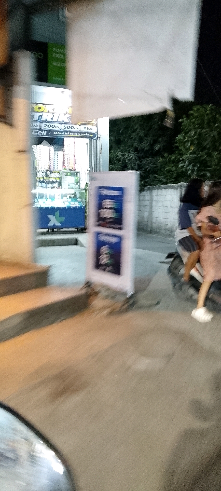 This is a blurry, low-light, outdoor image taken at night in the city of Satriajaya, Indonesia. In the foreground, a blurred motorcycle and two individuals are visible. One individual, wearing a light-colored dress and white shoes, is seated on the back of the motorcycle. The other individual, wearing a dark shirt and shorts, is operating the motorcycle. To the left, a blurred storefront is identifiable with illuminated signs and displays of merchandise, likely mobile phone accessories. A blue counter with an "X" logo is visible inside the store. A white poster with blue text is standing in front of the store. The background shows a brick wall and dense foliage. The lighting suggests artificial light sources from the storefront and streetlights.
