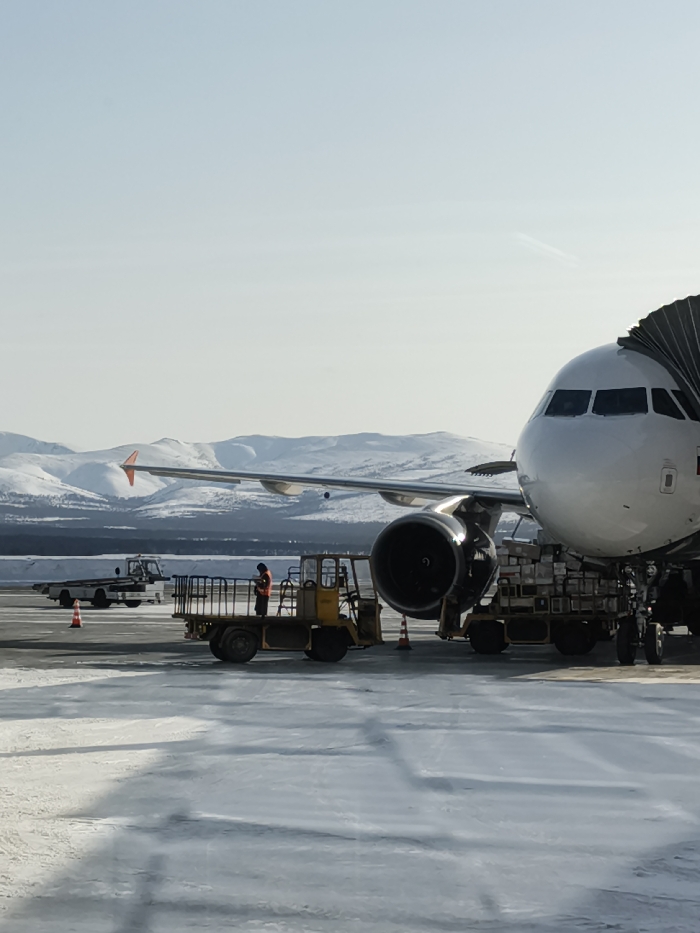 The image displays an active airport apron scene at Magadan Airport (Sokol Airport, UHMM) in Sokol, Magadan Oblast, Russia, under clear daylight conditions. A white commercial passenger aircraft, featuring a visible left wing, engine, and nose section, is parked at a gate with a black flexible jet bridge connected to its forward entry door. The aircraft displays a faint Russian flag decal on its fuselage near the cockpit windows, and the tip of its vertical stabilizer (rudder) appears orange/red.
The tarmac surface is partially covered with patches of snow and ice. In the mid-ground, a single individual wearing an orange high-visibility jacket is standing on the elevated platform of a yellow ground support cargo loader positioned near the aircraft's left engine. Several silver rectangular cargo containers, consistent with Unit Load Devices (ULDs), are stacked on multiple flatbed dollies directly beneath the aircraft's fuselage, indicating ongoing cargo loading or unloading operations. Further in the background, a white utility vehicle, possibly a tug, is parked on the apron to the left of the cargo loader. Several orange traffic cones are placed on the ground around the equipment. The background features a distinct range of snow-capped mountains under a clear, pale blue sky, with a dark line of trees visible at their base. A faint contrail is visible in the upper right sky.
