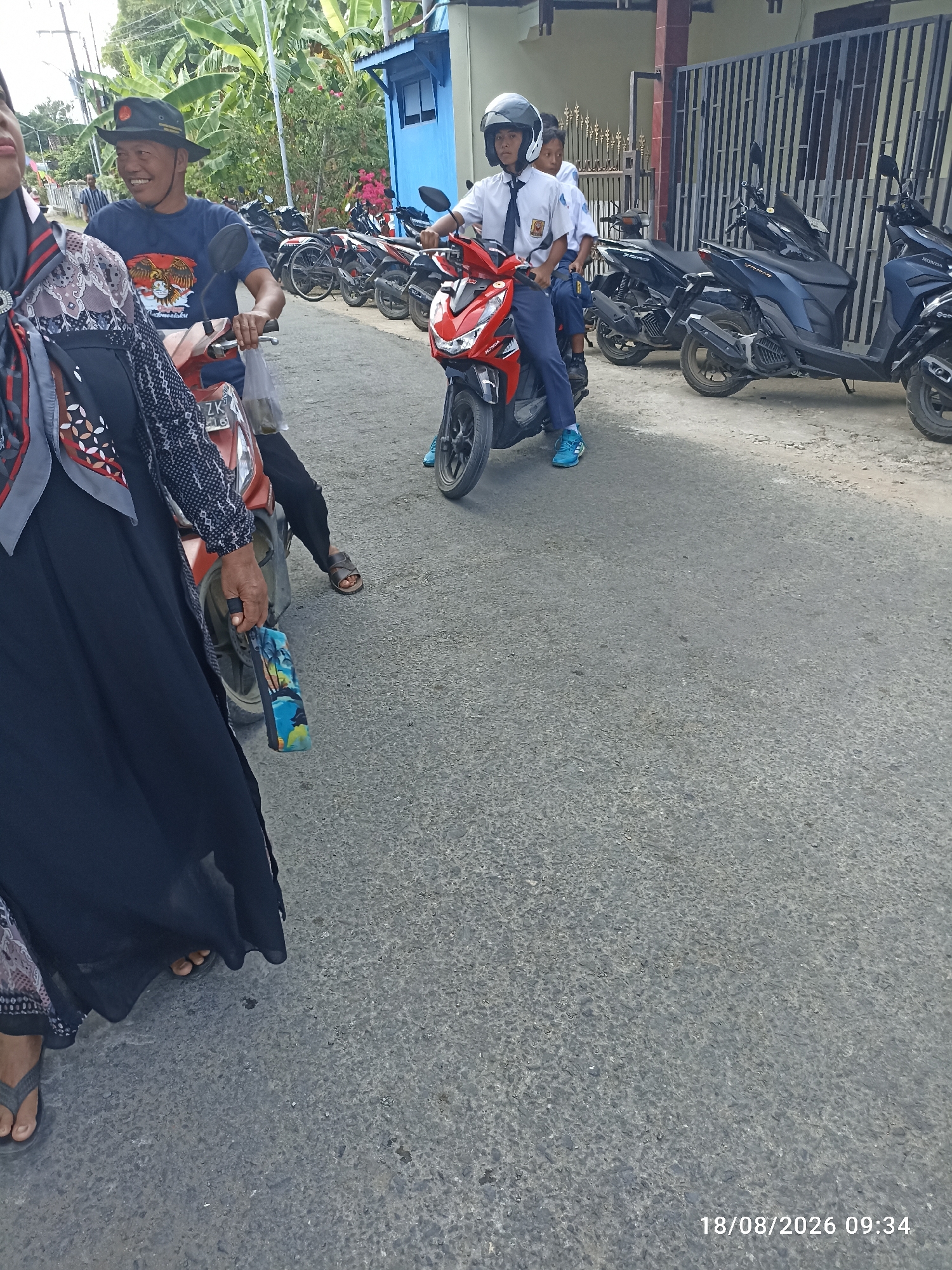 In Munjungan, Indonesia, on August 18, 2026, at 09:34 AM, a group of people are gathered near parked motorcycles on a paved road. In the foreground, a woman in a black abaya and headscarf stands next to a red scooter, holding a patterned bag. To her left, a man wearing a dark blue t-shirt and a wide-brimmed hat smiles as he sits on a scooter. Behind them, two young individuals in school uniforms and helmets ride on a red scooter. Several other motorcycles are parked along the roadside, with one particularly prominent dark blue scooter in the background. The environment appears to be a residential or communal area, with buildings and lush greenery visible.