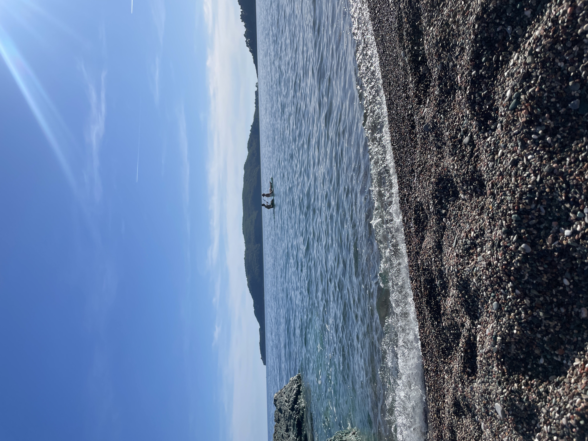 The image shows a beach in Rafailovići, Montenegro. The beach is covered in small, dark pebbles. The water is clear and blue, with small waves breaking on the shore. In the distance, there is a green, forested mountain. Two people can be seen paddleboarding on the water. The sky is blue with some white clouds and a contrail.