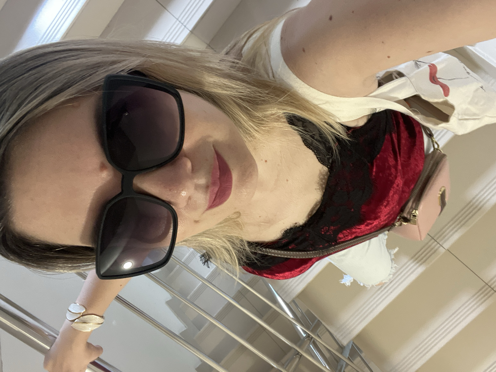 A woman is taking a selfie in a well-lit indoor setting, possibly a hotel or mall, with a staircase in the background. She has blonde hair and is wearing large, black sunglasses and bright pink lipstick. Her outfit consists of a white top over a red, velvet-like camisole with black lace trim. A small, light pink crossbody bag is visible. She is also wearing a distinctive gold and white cuff bracelet on her left wrist. The woman appears to be smiling or smirking, with a playful expression. The overall impression is of a casual yet stylish snapshot taken on a bright day. The stairs suggest an upscale or modern environment.