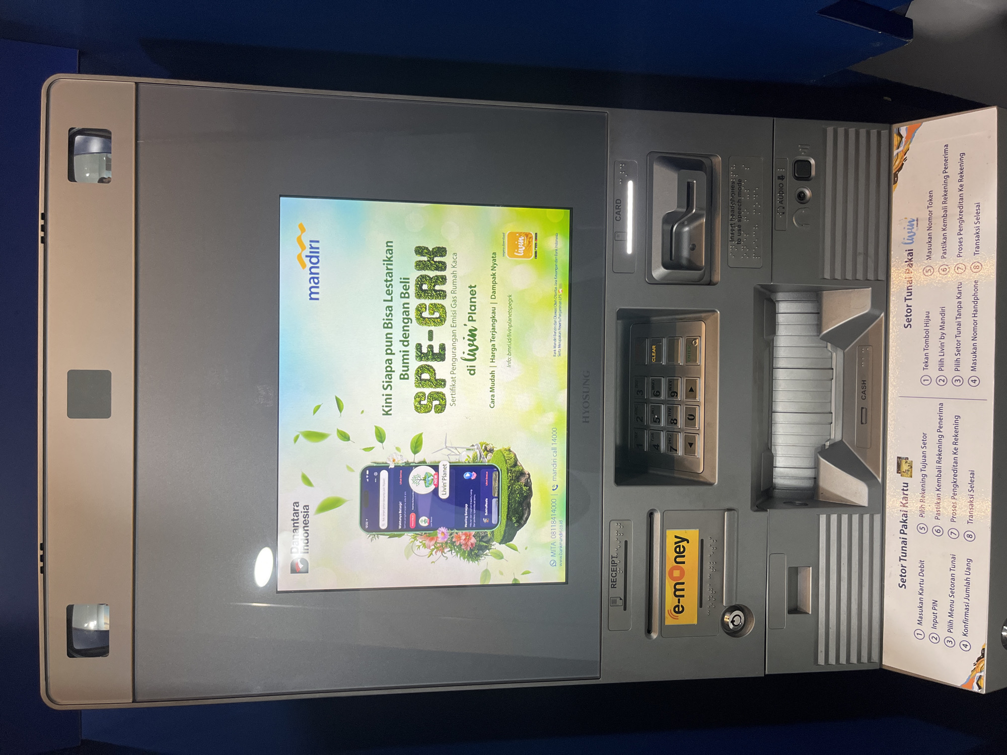 The image displays a modern, grey-colored ATM machine positioned indoors, against a blue background. No people are visible, and no specific activity is taking place.
The prominent central screen features an advertisement for "mandiri" bank, promoting an initiative called "SPE-GRK" (Sertifikat Pengurangan Emisi Gas Rumah Kaca) on their "livin' Planet" platform. The ad, written in Indonesian, translates to "Now Anyone Can Preserve Earth by Buying SPE-GRK (Greenhouse Gas Emission Reduction Certificate) on Livin' Planet. Easy Way | Affordable Price | Real Impact." It includes the "livin'" logo, "Danantara Indonesia" logo, and states that Bank Mandiri is licensed and supervised by OJK and Bank Indonesia. Contact information (Mita 08118414000, Mandiri Call 14000, www.bankmandiri.co.id) is also present. The ATM manufacturer "HYOSUNG" is visible on the screen's frame.
To the right of the screen are the ATM's functional components: a card slot labeled "CARD" with an illuminated entry, a cash deposit/dispensing slot labeled "CASH", and a numeric keypad featuring "CLEAR" and "ENTER" buttons. Below these, there's a receipt slot labeled "RECEIPT" and a yellow "e-money" slot. An audio jack is present, accompanied by text in English and Braille indicating "Insert headphones to use speech mode." A small security camera is located on the upper left of the machine.
On the far right, two instructional panels in Indonesian guide users on cash deposit procedures. One outlines "Setor Tunai Pakai Kartu" (Cash Deposit Using Card), detailing steps like inserting a debit card and inputting a PIN. The other, "Setor Tunai Pakai livin'", describes a cardless cash deposit using the "Livin' by Mandiri" app, involving pressing a green button and entering a phone and token number. Both procedures conclude with confirming the recipient account and completing the transaction. The use of Indonesian language and Mandiri bank branding indicates the location is in Indonesia, specifically West Jakarta.