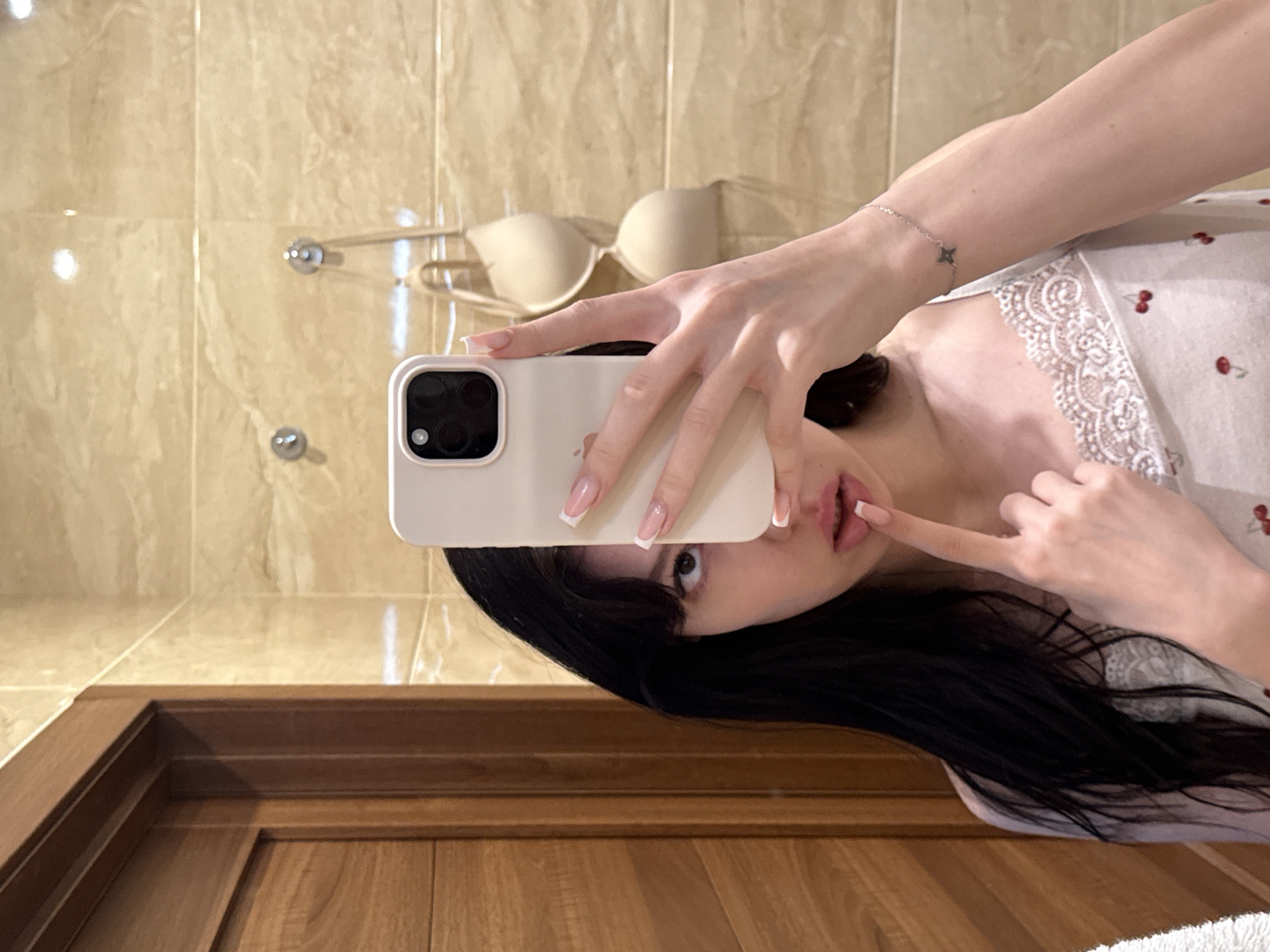 A woman with long, dark hair and light skin is taking a selfie in a bathroom. She is wearing a white top with red cherry patterns and lace trim. Her left hand is holding a phone with a white case, and her right hand is touching her lips. In the background, beige tiles cover the wall, and a wooden door is visible on the left. Two nude bras are hanging on hooks on the tiled wall. The scene appears to be indoors, possibly in an apartment in Khimki, Russia.