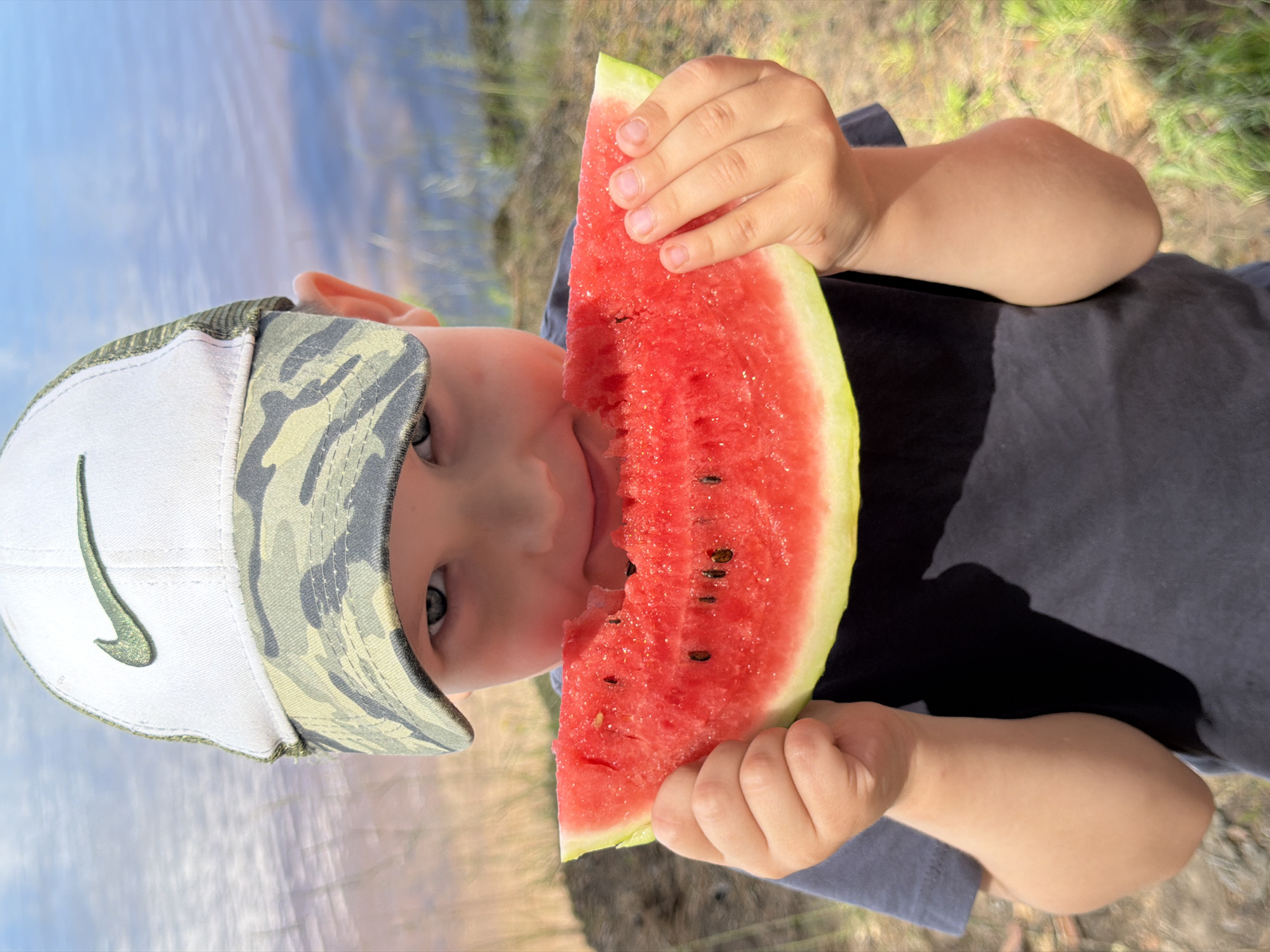 A young boy wearing a camouflage-patterned baseball cap with a green Nike logo is holding a slice of watermelon and eating it. He is outdoors with a body of water and vegetation in the background. The lighting suggests it is daytime. The scene does not provide any specific visual cues to identify the location as Chaikovsky, Russia.