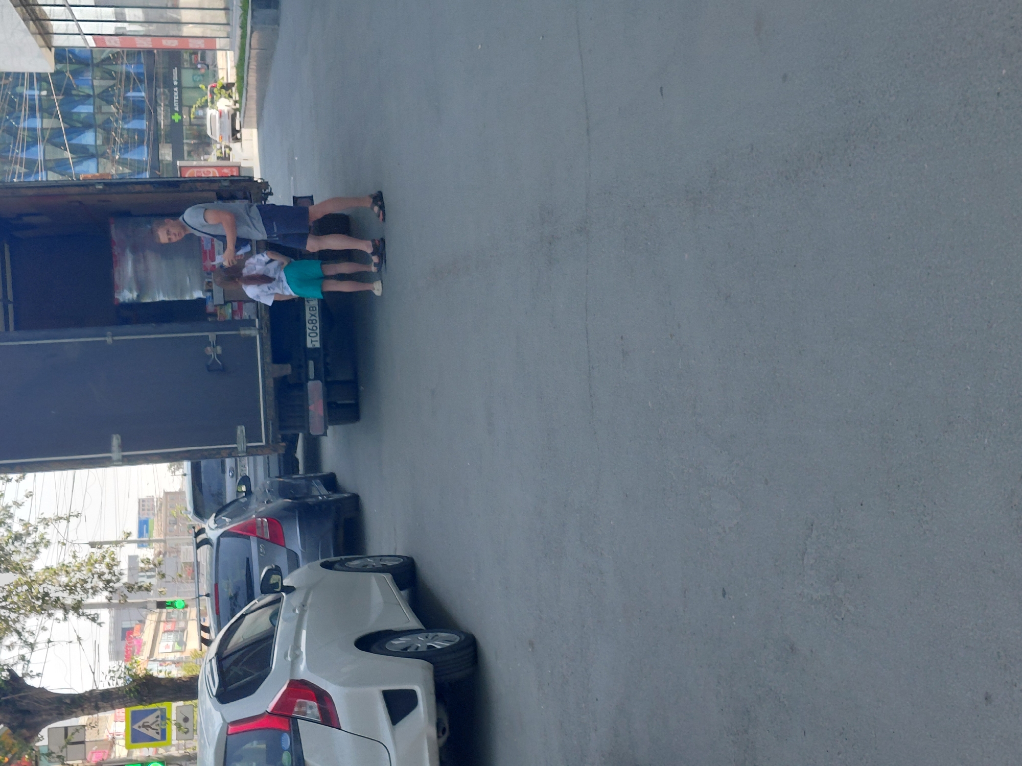 The image depicts a street scene in Novosibirsk, Russia, likely during daytime. In the foreground, the asphalt road is visible, with a white Honda Fit parked on the left. Further down the street, a gray hatchback is also parked. A large truck, with its rear doors open, occupies a significant portion of the right side of the frame.
Two individuals are interacting near the open truck. A man, dressed in a gray t-shirt and blue shorts, stands facing away from the viewer, holding what appears to be a box or package. A young girl, wearing a white t-shirt and teal shorts, stands to his left, also facing away from the viewer. She appears to be holding something small in her hands. The truck's license plate is partially visible, showing "T068XB1".
In the background, across the street, a modern building with a glass facade is visible, along with other structures and streetlights. Trees with sparse foliage are present on the left side of the image, suggesting a season other than peak summer. Pedestrian crossing signs and traffic lights are also visible, indicating an urban environment with traffic management. The overall impression is one of everyday urban activity, possibly related to delivery or unloading from the truck.