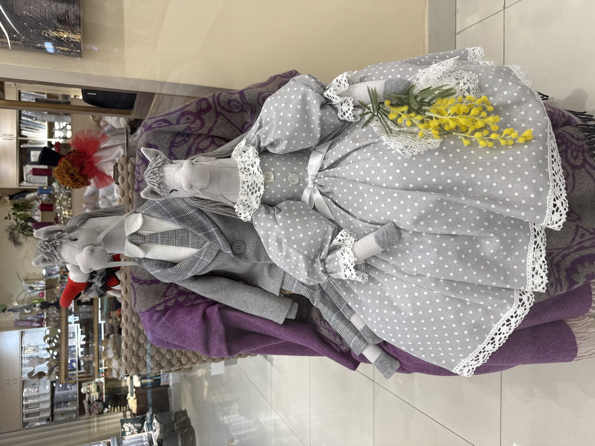 This is a medium shot of two handmade plush horse dolls, dressed in formal attire, positioned on a plush chair. The horse on the left wears a gray plaid suit with a white shirt and a gray tie. The horse on the right is adorned in a gray polka-dotted dress with puffy sleeves, a lace collar, and a white ribbon sash. The dolls are seated on a chair covered with a purple throw blanket and a gray patterned blanket. In the background, shelves are filled with various items, suggesting a retail environment. A branch of yellow flowers is tucked into the dress of the horse on the right. The floor appears to be tiled. The overall scene is indoors, likely in a shop or market. The image itself was taken on March 10, 2023. The location is a shop in Izhevsk, Russia.