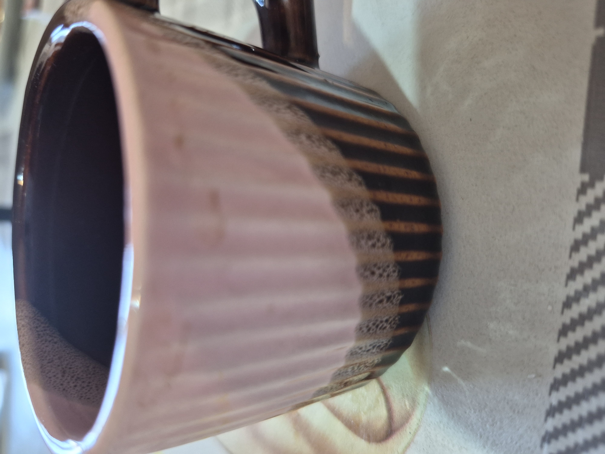A close-up, rotated shot captures a two-toned ceramic mug resting on a textured surface. The mug features a glossy, light brown or muted pink upper section that transitions to a dark, rich brown base with distinct horizontal ribs or grooves. At the border where the two colors meet, there's a visible textural detail, possibly from the glaze application, appearing slightly bubbly or frothy. The mug's handle, partially visible in the upper right, is also dark brown. The interior of the mug is a deep, dark brown.
The surface beneath the mug is a light, possibly off-white or very light grey, with a subtle grainy texture. To the right, a segment of a broader pattern is visible, featuring alternating dark grey and white chevron stripes, suggesting a tablecloth or mat. The lighting is bright, creating reflective highlights on the mug's glazed surface, particularly along its rim and handle, indicating an indoor setting. No people, discernible activities, or specific location cues are visible within the frame. There is no visible text.