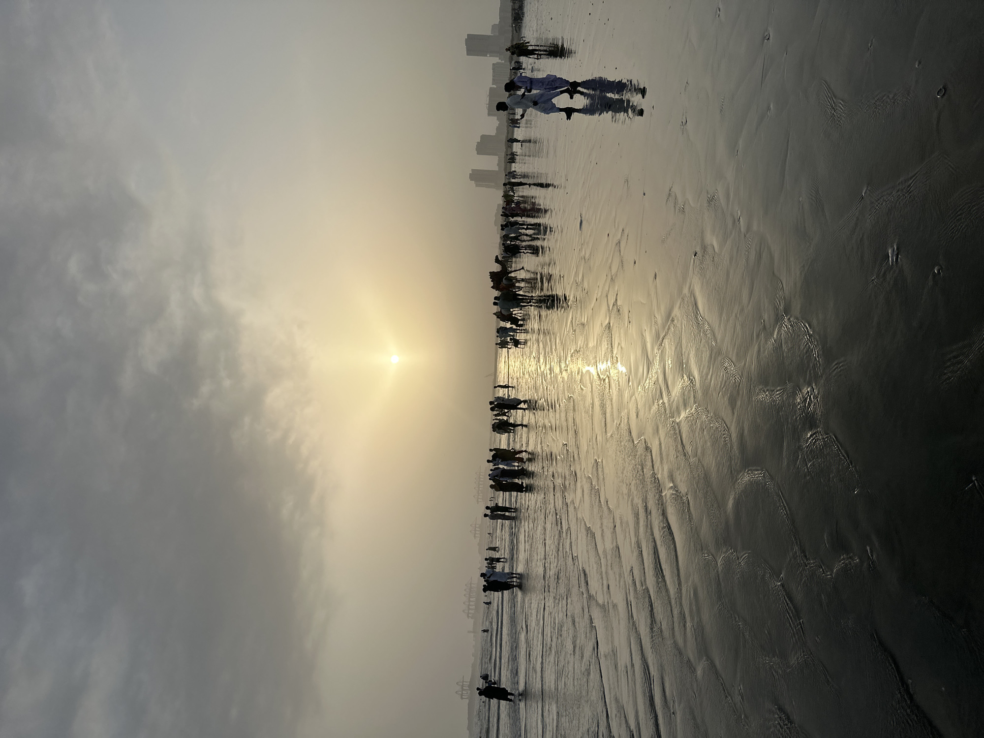 The image depicts a beach scene during sunset, with people walking along the wet sand. The sun is low in the sky, casting a warm glow and creating reflections on the water. Silhouetted figures of people are visible, some walking alone, others in groups. In the distance, the outline of a city skyline can be seen, suggesting an urban coastal setting. The clouds are diffused, with the sun breaking through them, indicating hazy or overcast conditions. The sand shows ripples and patterns from receding waves, adding texture to the foreground. The overall mood is tranquil and serene, capturing the beauty of a sunset at the beach. The orientation of the image is vertical.