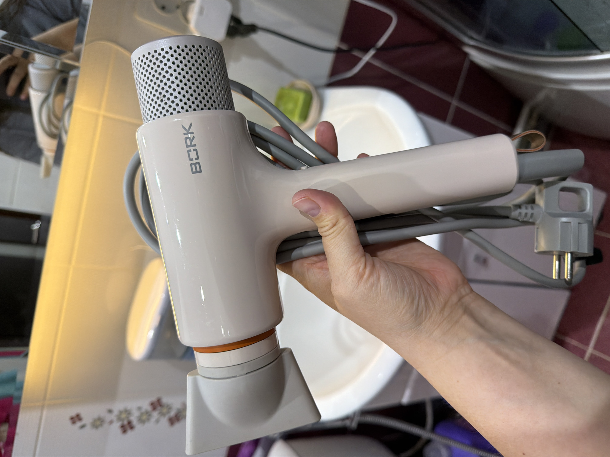A hand holds a Bork hair dryer against a background that suggests a bathroom setting. The hair dryer is mostly pale beige with a perforated silver cylinder at the top and an orange ring where the nozzle attachment connects. The grey power cord is wrapped around the hair dryer and terminates in a European plug. The background features a white sink or basin and wall tiles. The image was captured indoors. The location is not discernible as Chaikovsky, Russia.