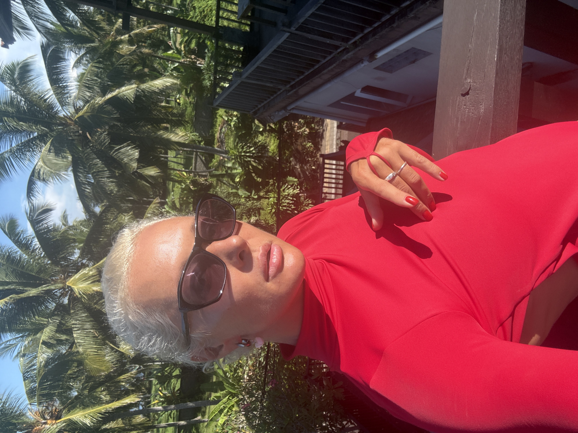 A woman with short, white hair and sunglasses is wearing a red long-sleeved dress. She is standing outside with palm trees and other lush greenery in the background. A building with a dark wooden balcony is partially visible. The location is Unknown, Unknown.