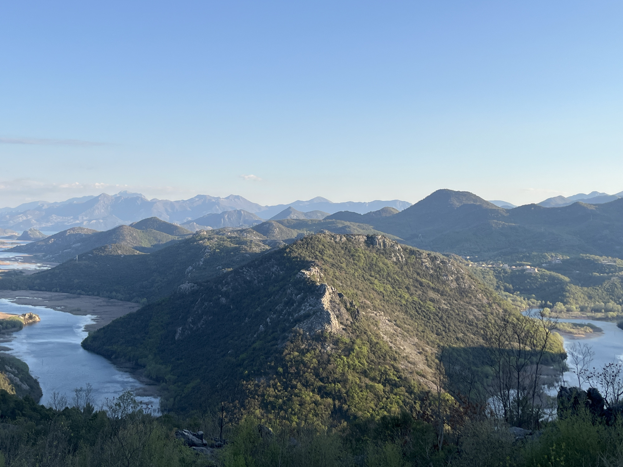 The image shows a wide panoramic view of the landscape in Meterizi, Montenegro. The foreground consists of wooded vegetation with branches extending into the sky. A river or large body of water with reed beds extends from the left side of the image and snakes between green, mountainous terrain. Mountain ranges are visible in the mid-ground, with some having exposed rocky terrain. In the background, layers of distant, blueish mountain ranges meet a clear blue sky. There are no people visible in the frame.