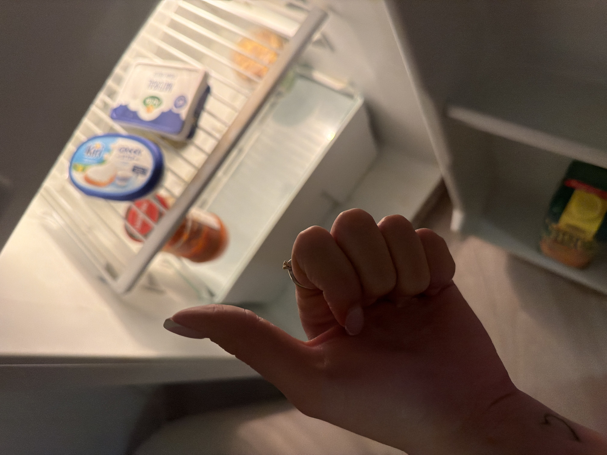The image displays a close-up view looking into an open refrigerator, dominated by a human hand making a "thumbs up" gesture. The hand, which has light-colored manicured fingernails, wears a simple gold-colored ring with a small stone on the ring finger. A faint, dark, wave-like mark or tattoo is visible on the person's wrist in one of the cropped views.
Inside the well-lit refrigerator, multiple shelves are visible. On the middle shelf, two distinct food items are present. One is an oval-shaped light blue container with the text "Kiri Greek" clearly visible on its label. Just behind it and slightly to the right, a rectangular white package with purple branding can be seen, with the word "Natural" printed upside down on its label. A round, red and white patterned object, possibly a snack or condiment, is also on this shelf. On an upper shelf in the background, a glass jar with a dark green label and a yellow lid is partially visible. The overall lighting suggests an indoor setting, likely in a dimly lit room where the refrigerator's internal light is the primary illumination source.