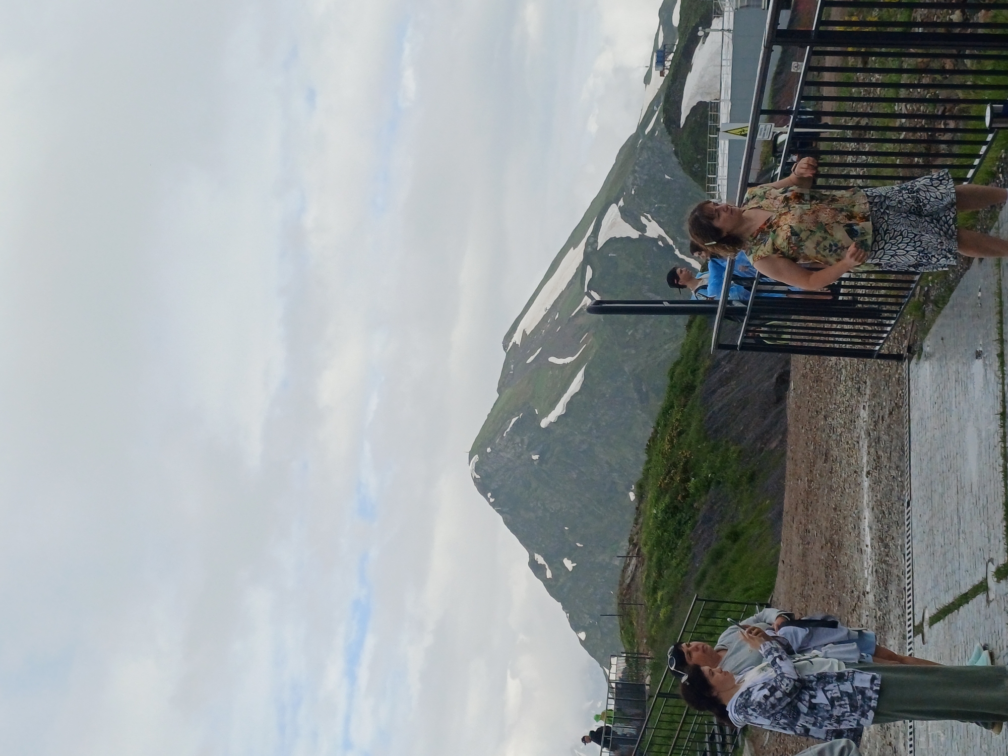 In Estosadok, Russia, a group of people are gathered on an elevated walkway overlooking a mountain landscape. The mountain is partially covered in snow, suggesting a cooler climate or high altitude. The sky is overcast with clouds, indicating a potentially moody or damp day.
One woman, wearing a floral print top and a patterned skirt, leans against a black metal railing, her head tilted back as if enjoying the view or the moment. Nearby, another person in a blue shirt stands, also looking out at the scenery. Further along the walkway, two more individuals are visible. One person, in a grey sweater and shorts, is looking at their phone, while another, wearing a patterned shirt and a long skirt, is also engaged with a device. They are all situated on a paved walkway, with a steep, grassy slope rising behind them and the impressive mountain face dominating the background. The overall scene depicts a leisurely moment of sightseeing amidst a beautiful, natural environment. There is no visible text in the image.