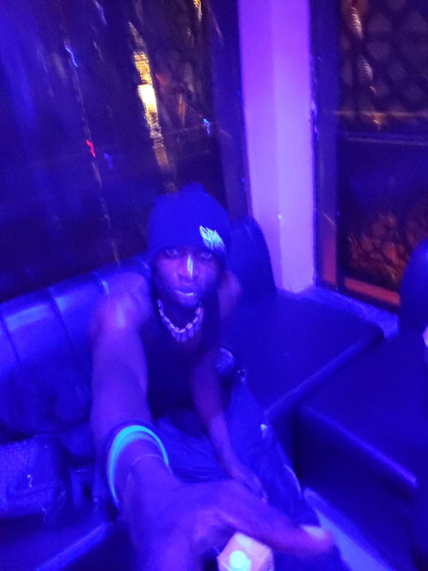 The image shows a person wearing a black beanie and a black tank top, sitting on a dark couch under blue and purple lighting. The person has white markings on their face, possibly from face paint. They are also wearing a necklace with light-colored beads and a neon green bracelet on their left wrist. The background is dark with some blurry lights and reflections, suggesting an indoor environment possibly a club or a party. The lighting creates a vibrant, almost neon effect. There is no visible indication of the city of Jos, Nigeria, in the image.