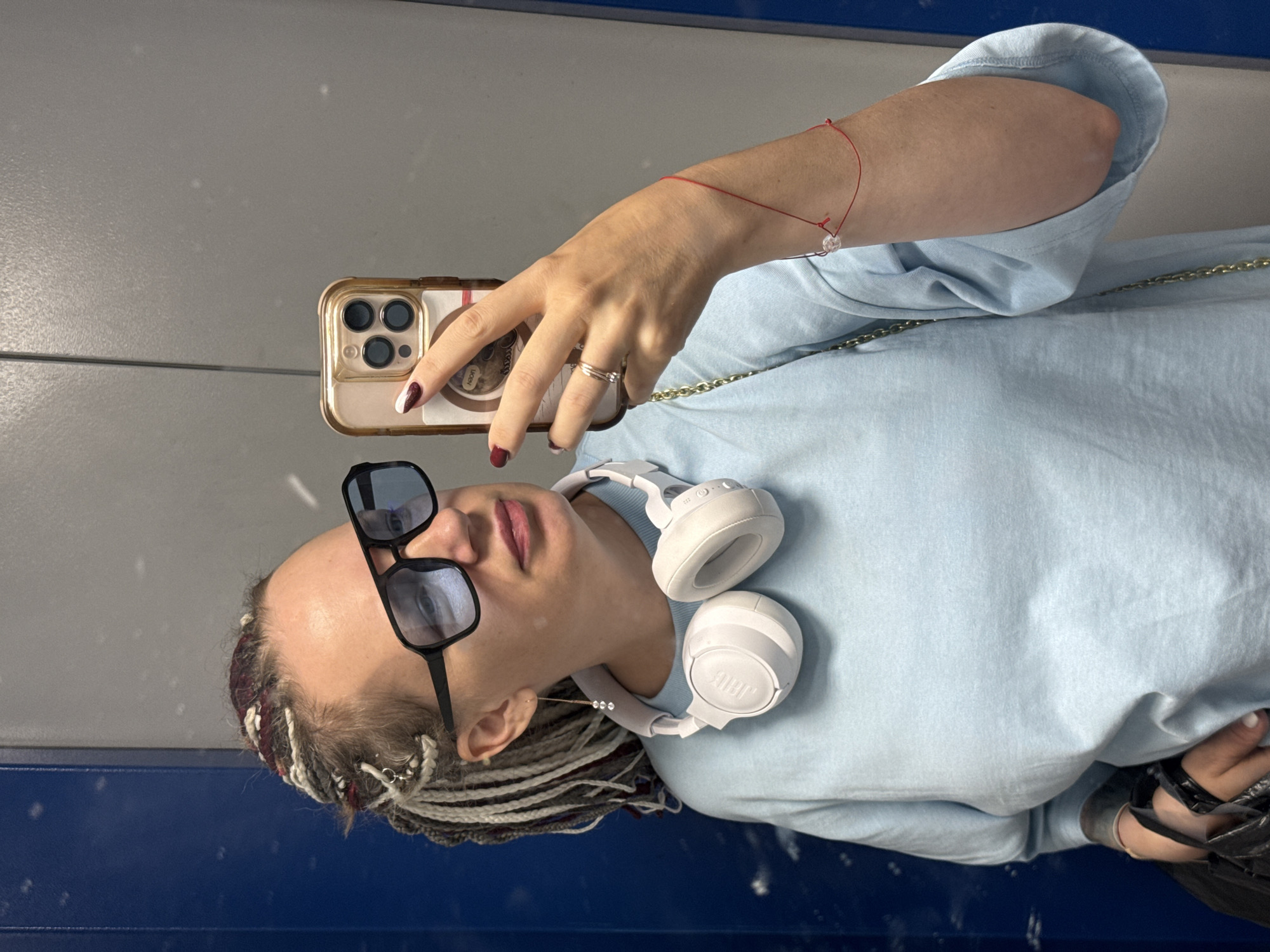 A person is taking a selfie in a dimly lit indoor environment, possibly a train or subway car, given the blue and grey paneling visible in the background. The person, seen from the chest up, is wearing a light blue long-sleeved shirt and white headphones around their neck. They are also wearing large black-rimmed sunglasses and have their hair in braided style with streaks of white and red. A thin red string bracelet is on their left wrist, and they are wearing a silver ring on their left ring finger. The person is holding a smartphone in their right hand, angled to capture the reflection in what appears to be a mirror or polished surface. Their fingernails are painted a deep red with white tips. The overall mood is casual. No specific time of day or weather conditions are discernible. There is no visible text in the image.