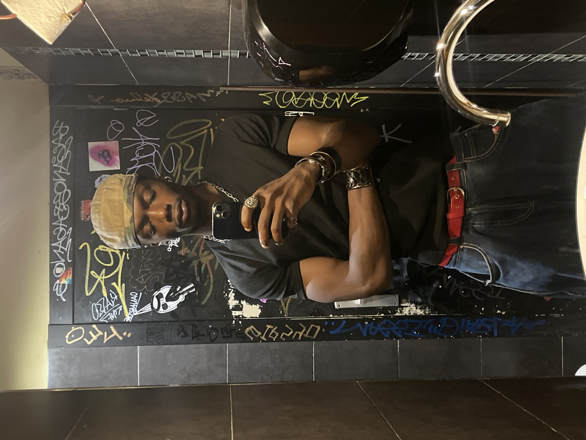 The image shows a person taking a selfie in a bathroom. The person is wearing a black t-shirt, blue jeans, a red belt, and a camouflage cap. They are also wearing several rings and bracelets. The bathroom has graffiti on the walls and a chrome faucet is visible in the foreground. The person's reflection is visible in the mirror. The image does not provide any information about the location in the city of Barcelona, Spain.