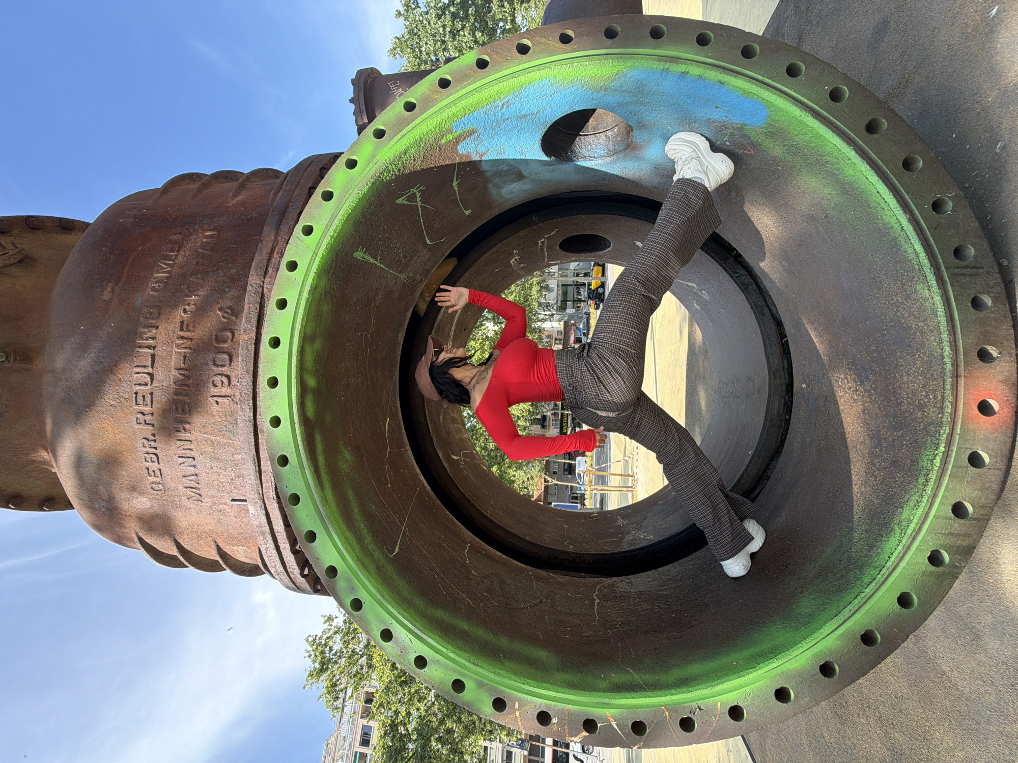 A woman in a red long-sleeved shirt, brown plaid pants, and white sneakers is striking a pose inside a large, circular industrial valve, located in Barcelona, Spain. The valve is constructed of rusty metal with a green and blue painted interior ring. The woman is positioned horizontally within the valve, with one foot resting on the inside wall and her hand reaching out. The background shows trees, buildings, and blue sky. The valve has text and numbers imprinted on the rusty metal.