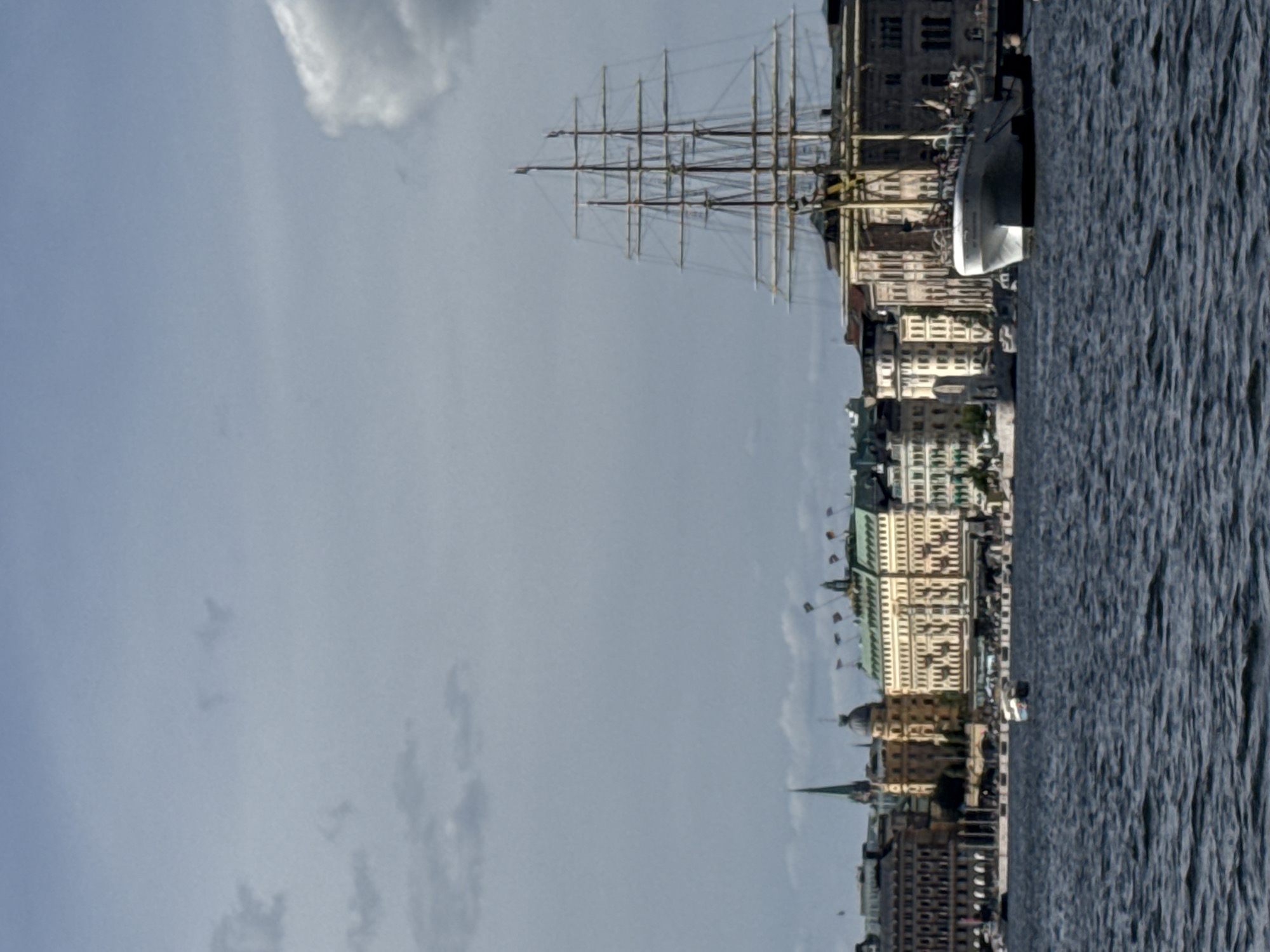 A wide-angle, full-color, horizontal photograph shows a city skyline with a large sailboat docked in the foreground. The sky is a pale blue with some scattered clouds. The water in the foreground is dark blue and choppy.
The sailboat is a tall ship with multiple masts and rigging. It is white with a yellow stripe along its hull. People can be seen on the deck of the sailboat.
In the background is a city skyline with several buildings. The buildings are of various heights and architectural styles, with some appearing to be older with ornate facades, while others are more modern. Flags are visible on some of the buildings. The skyline suggests an urban setting, possibly a harbor or waterfront area.
The overall impression is a picturesque scene of a historic city with a prominent sailing vessel. The location is identified as Stockholm, Sweden, a city known for its archipelago and maritime history. The specific buildings in the background are consistent with the architecture found along the waterfronts of Stockholm.