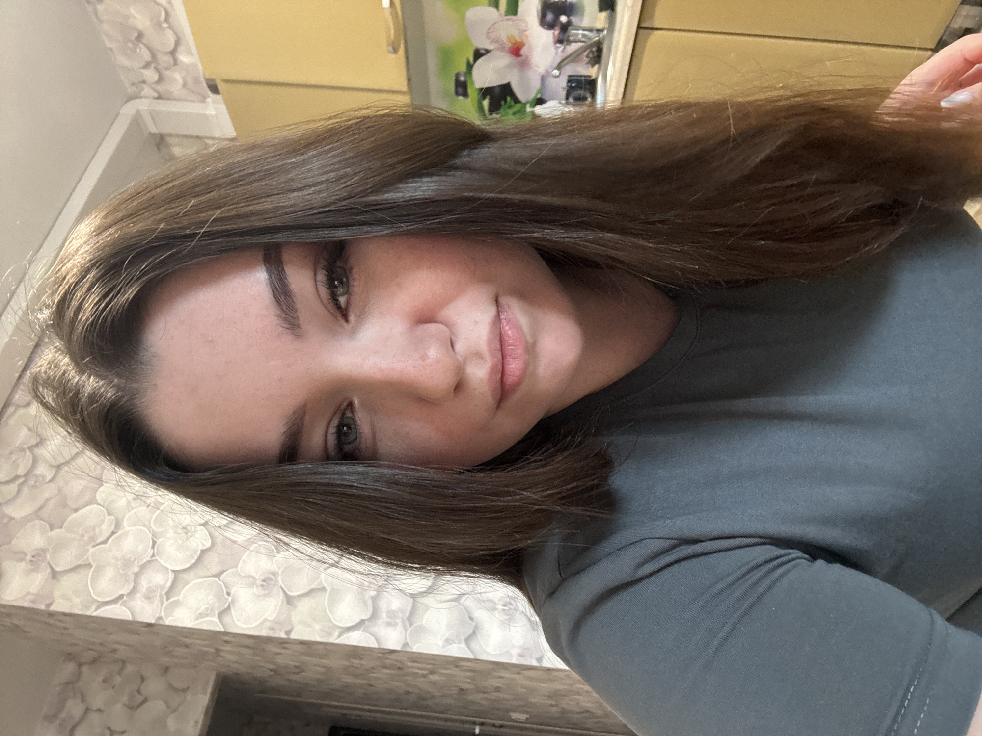 A close-up, head-and-shoulders selfie shows a young woman with long, dark brown hair partially obscuring her face. She has light-colored eyes, dark eyebrows, and fair skin with freckles. She is wearing a dark gray t-shirt. The background is out of focus and appears to be an indoor setting with patterned wallpaper on the left and light yellow kitchen cabinets on the right. A sink and a picture of an orchid are visible on the kitchen counter. The lighting suggests an interior space with natural light coming from the left. The scene is located in Zarechnaya (Gamovskoe S/P), Russia.