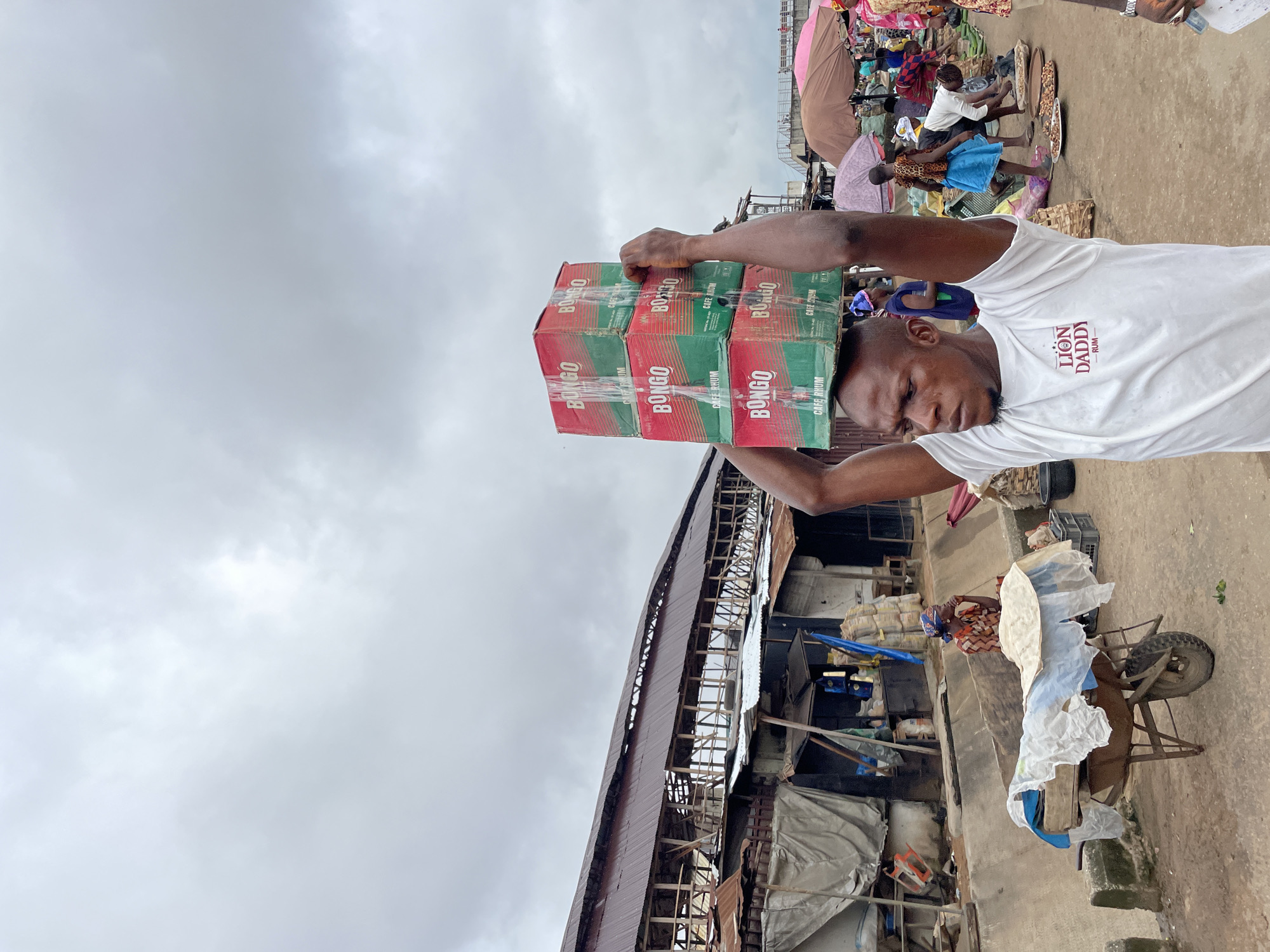 This is a vertical, eye-level photograph taken in a busy outdoor market, likely in Onitsha, Nigeria. The main subject is a man wearing a white t-shirt with "LION DADDY RUM" printed on it. He is carrying a stack of six red and green cardboard boxes, labeled "BONGO CAFE RHUM," balanced on his head. His head is tilted forward, and his facial expression suggests exertion.
In the background, the market scene unfolds. Several people are visible, some standing and interacting, while others are seated. There are market stalls with goods displayed, and various objects including bags, baskets, and what appears to be a wheelbarrow with a person nearby. The structures in the background are rudimentary, with corrugated iron roofs and open sides, typical of many African markets. The ground is dusty and uneven.
The sky is overcast with heavy clouds, suggesting it might be a humid or rainy day. The lighting is diffused, indicating it is likely daytime, possibly late morning or afternoon. The overall impression is of a vibrant, working market environment where people are engaged in commerce and transportation of goods. The visible text on the boxes and the man's shirt provides brand information, adding a layer of commercial context to the scene.