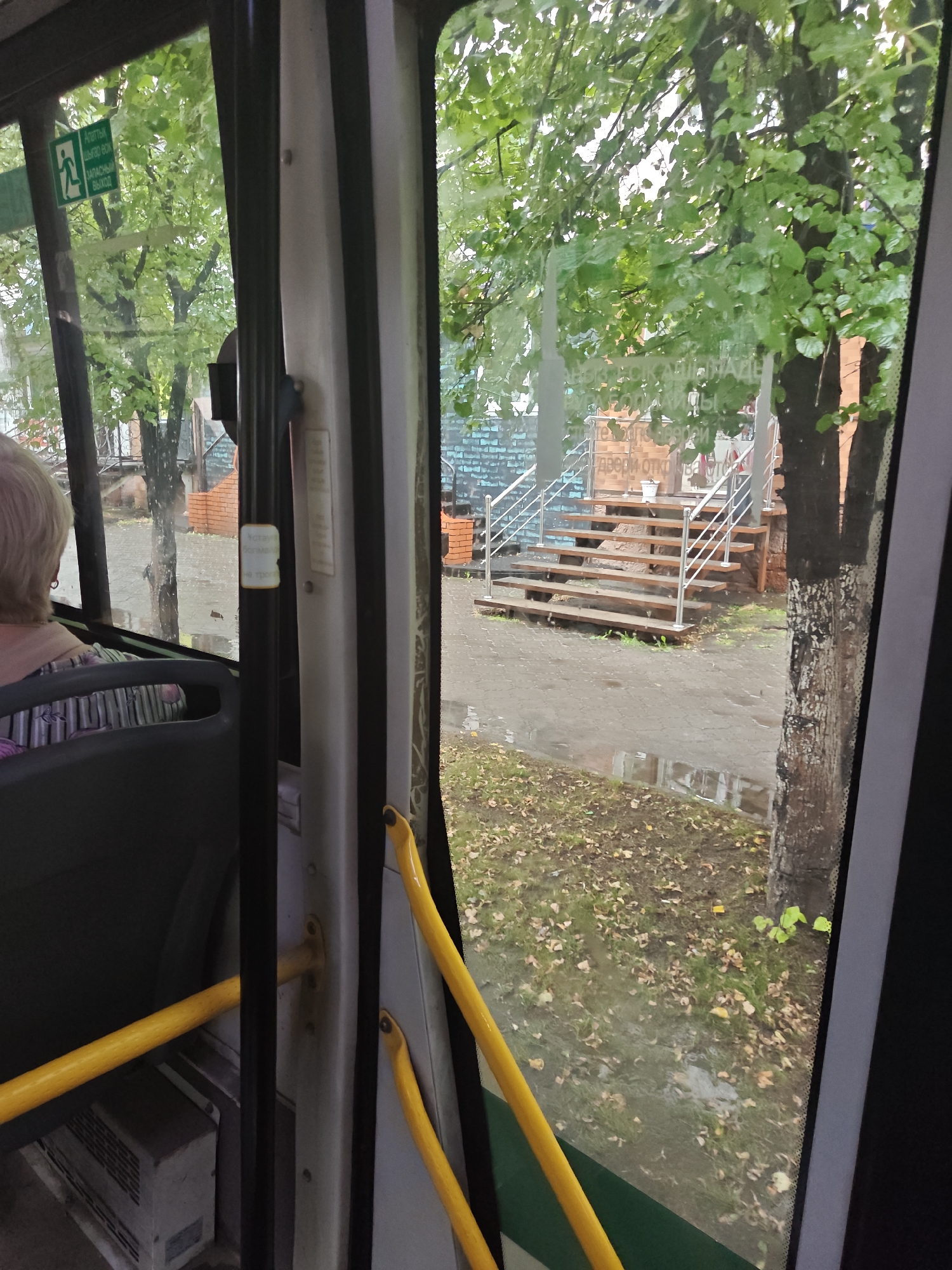 The image was taken from inside a bus in Petropavl, Kazakhstan. Through the window, we see a staircase with metal railings leading up to a building with brick and blue tiled walls. The ground is wet and there are fallen leaves on the grass and pavement, indicating it may have recently rained. The bus has yellow grab bars inside. A woman with blonde hair is seated on the bus. A green sign with a white pictogram of a person running out of a door is visible on the window.