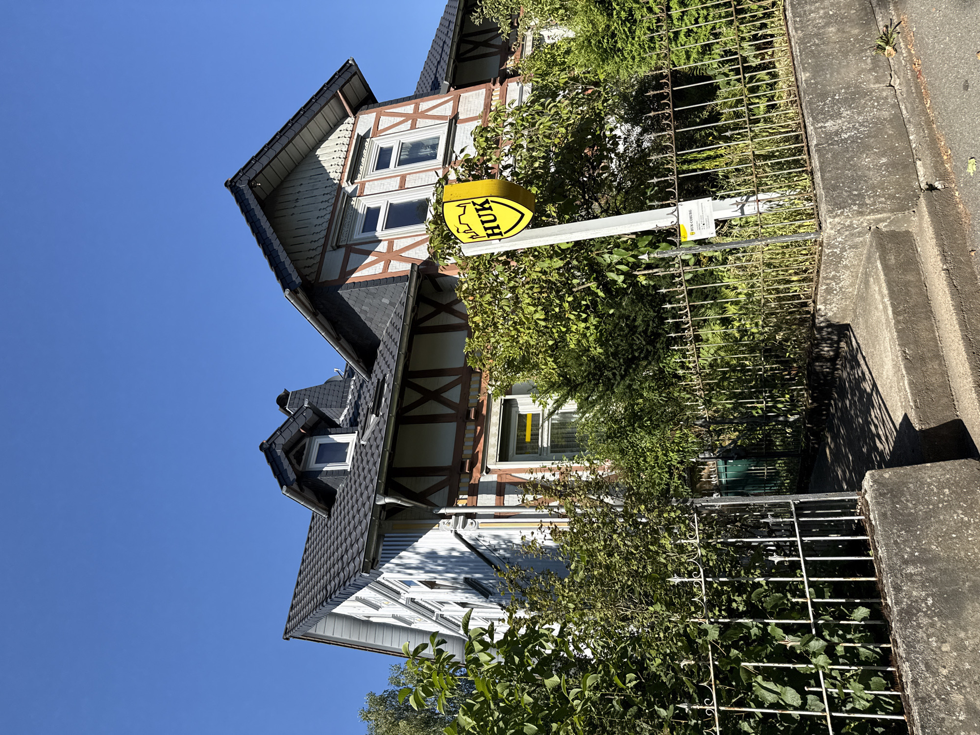 The image shows a two-story building with a dark tiled roof and white exterior walls, featuring half-timbered accents in brown and white. The building has several windows, some with white frames and others with darker frames, and a prominent dormer window on the upper level. In front of the building, there is a white metal fence and dense green foliage, including trees and bushes. A yellow sign with a shield emblem and the text "HUK" is mounted on a pole in front of the fence. The ground in front of the fence is a paved walkway leading up to the property. The sky is clear and blue. The scene appears to be on a sunny day in a residential or commercial area. The building's architectural style suggests it is located in Germany, and the presence of the "HUK" sign, which is a German insurance company, further supports this. A sign at the bottom of the pole indicates the company is HUK-COBURG. The location is identified as Osterode am Harz, Germany.