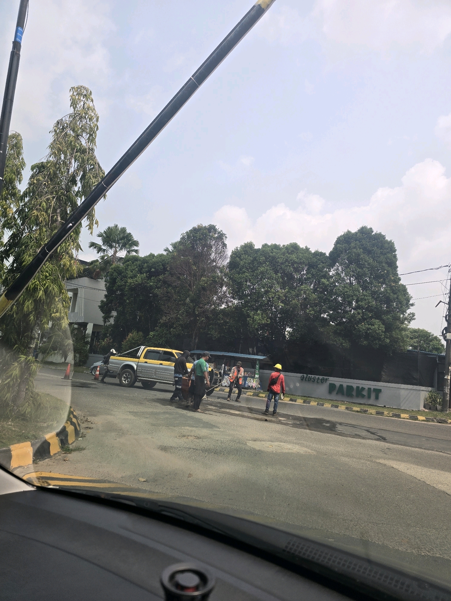 In Batam, Indonesia, on a street with a paved road and a yellow and black striped curb, several individuals are present. A yellow and silver pickup truck is parked on the right side of the road. Near the truck, a group of people, some wearing high-visibility vests, appear to be engaged in an activity, possibly related to work or assistance.
Further down the road, a building with the signage "cluster PARKIT" is visible. Two individuals, one wearing an orange vest and the other a red shirt and yellow hard hat, stand near the building. The environment is characterized by trees, including palm trees, and a cloudy sky overhead. The view appears to be from inside a vehicle, indicated by the car's dashboard and windshield in the foreground.