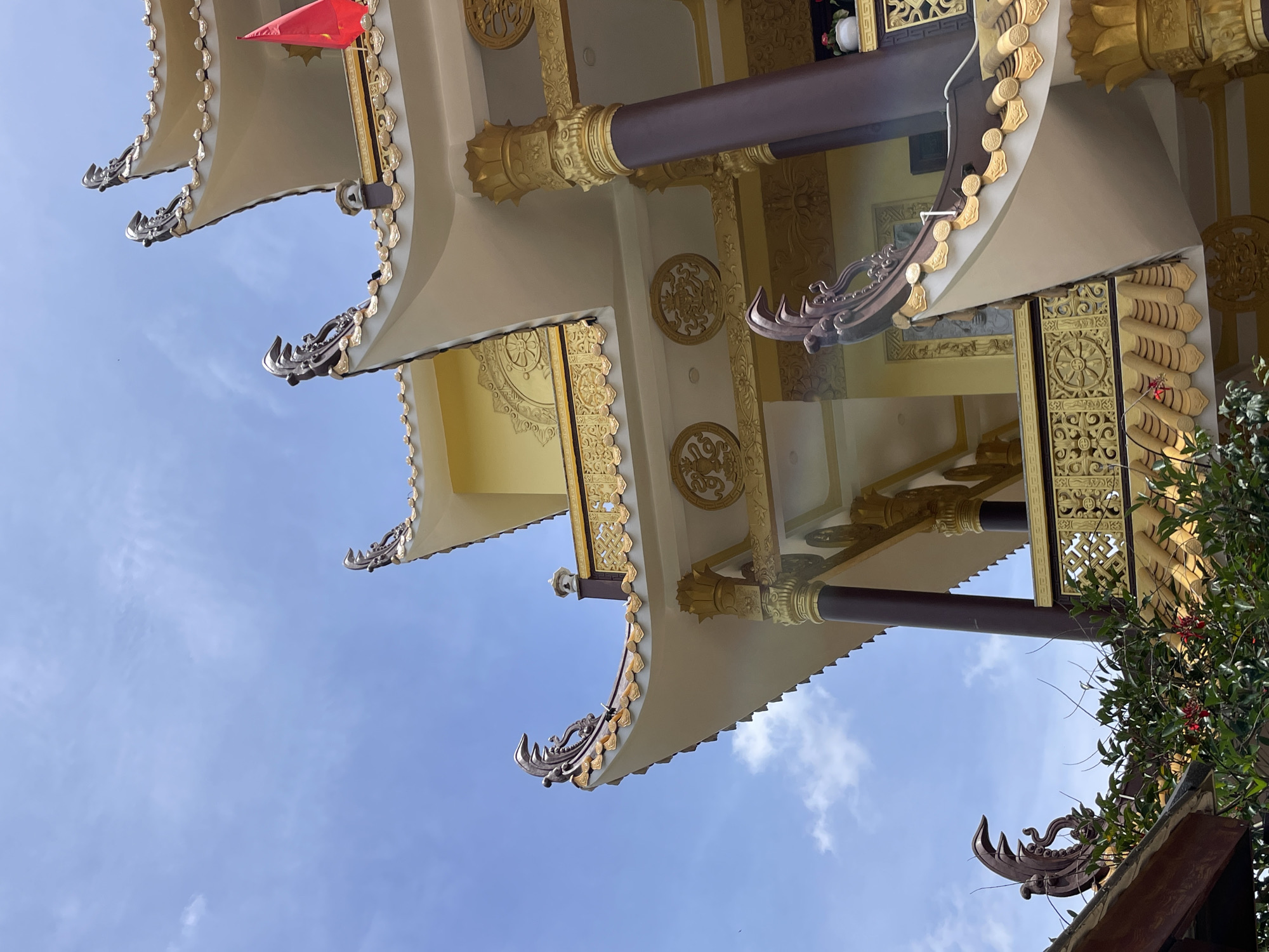 The image shows a close-up, low-angle view of a pagoda roof against a partly cloudy blue sky. The pagoda features traditional East Asian architectural elements, including curved eaves decorated with golden trim and ornate, dark brown finials on the corners. The roof itself is tiled in a golden hue, and the building's walls are painted yellow. Decorative elements in gold, such as circular motifs with intricate designs and patterned railings, are visible. A vibrant red flag is attached to the top of the pagoda. Lush green foliage with small red flowers is in the foreground, partially obscuring the lower levels of the structure. The scene appears to be from Truc Lam Zen Monastery in Da Lat, Vietnam, a prominent Buddhist temple known for its architecture and scenic location. There are no people visible in the image.